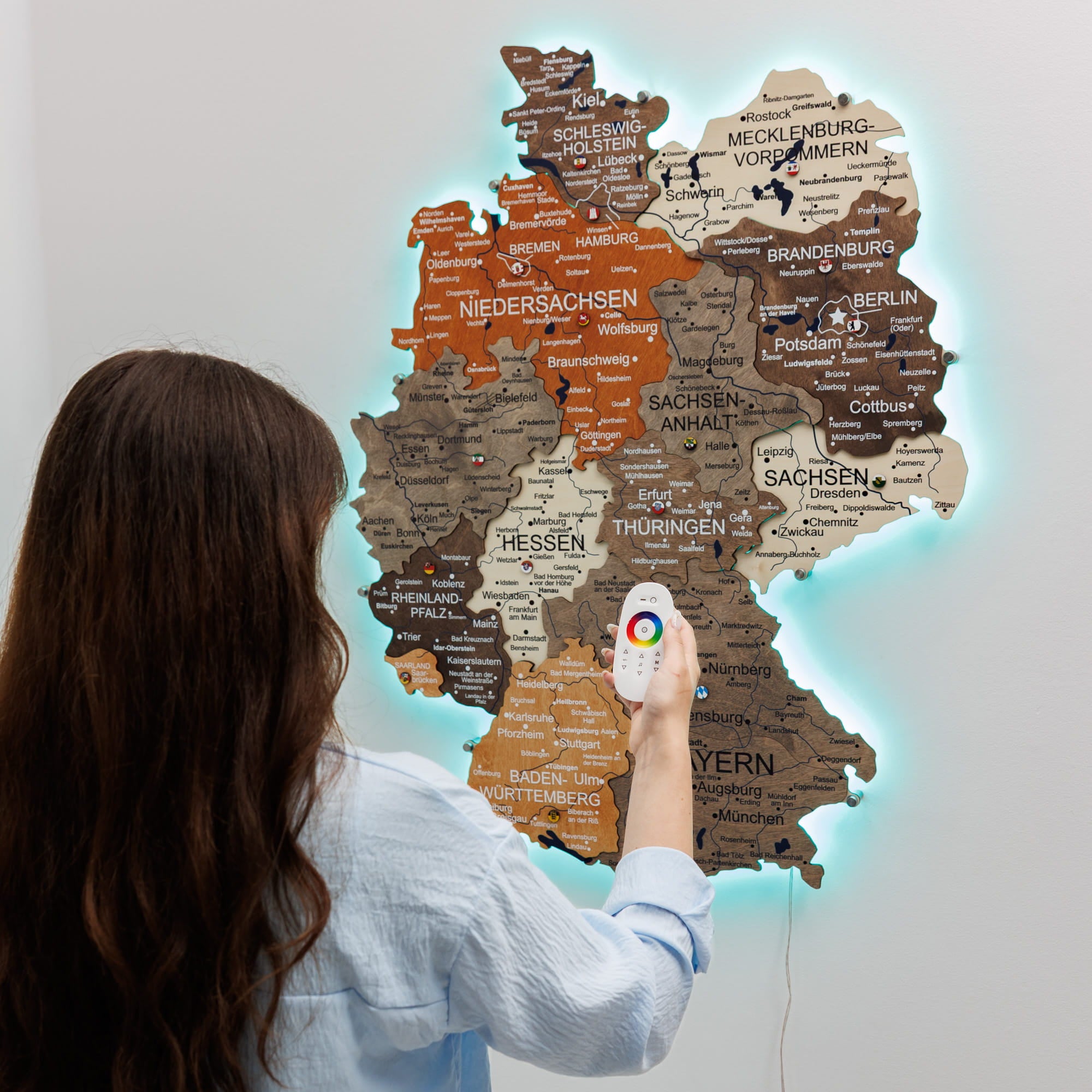 A woman stands in front of a glowing, illuminated map wall of Germany, holding a white remote control with a colorful dial. The map is detailed with labels for various German states and cities, and glows with a bright cyan light against a plain light gray wall.