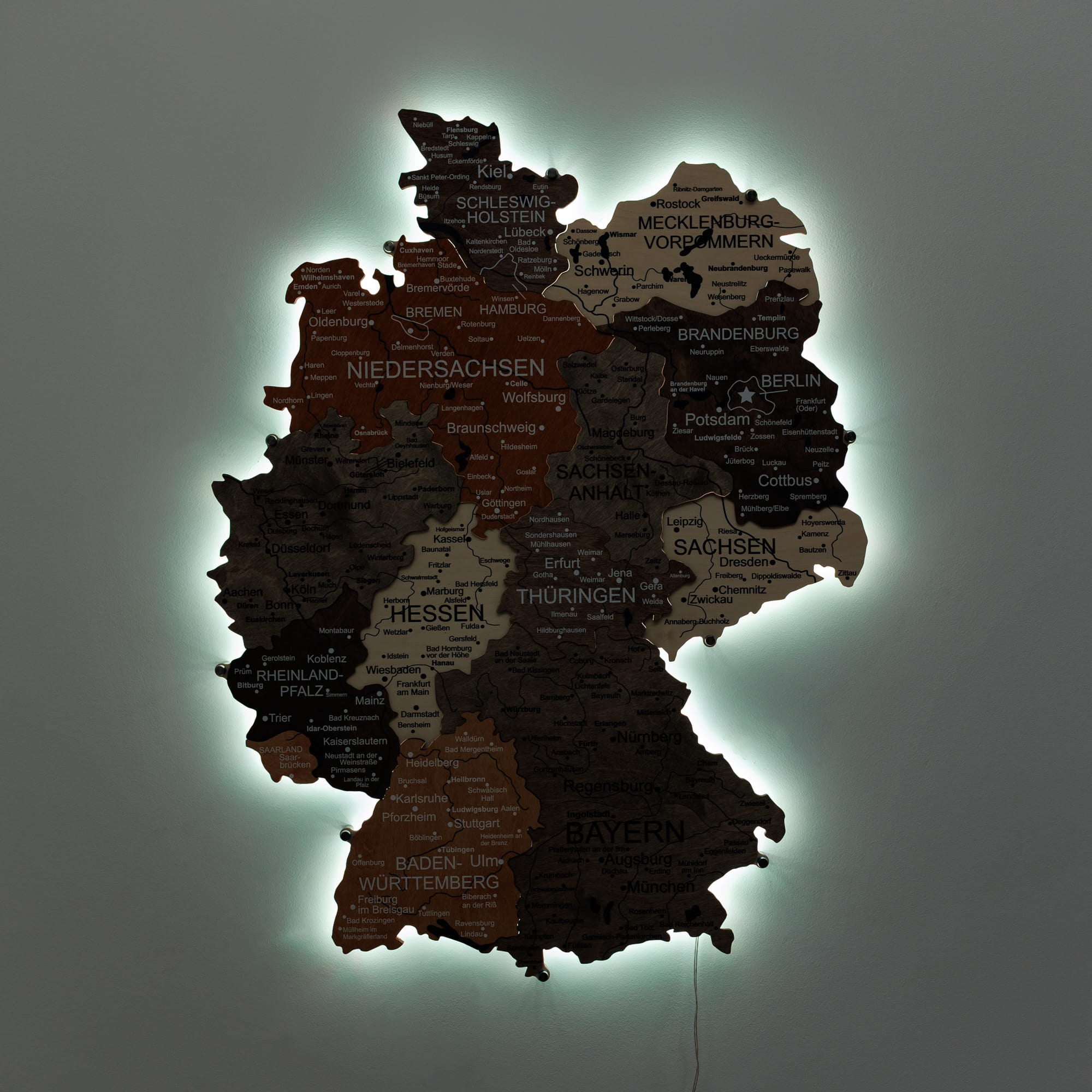 A 3D illuminated wall map of Germany, showing states and cities in dark brown, black, and beige tones with a glowing green backlight against a dark gray background.