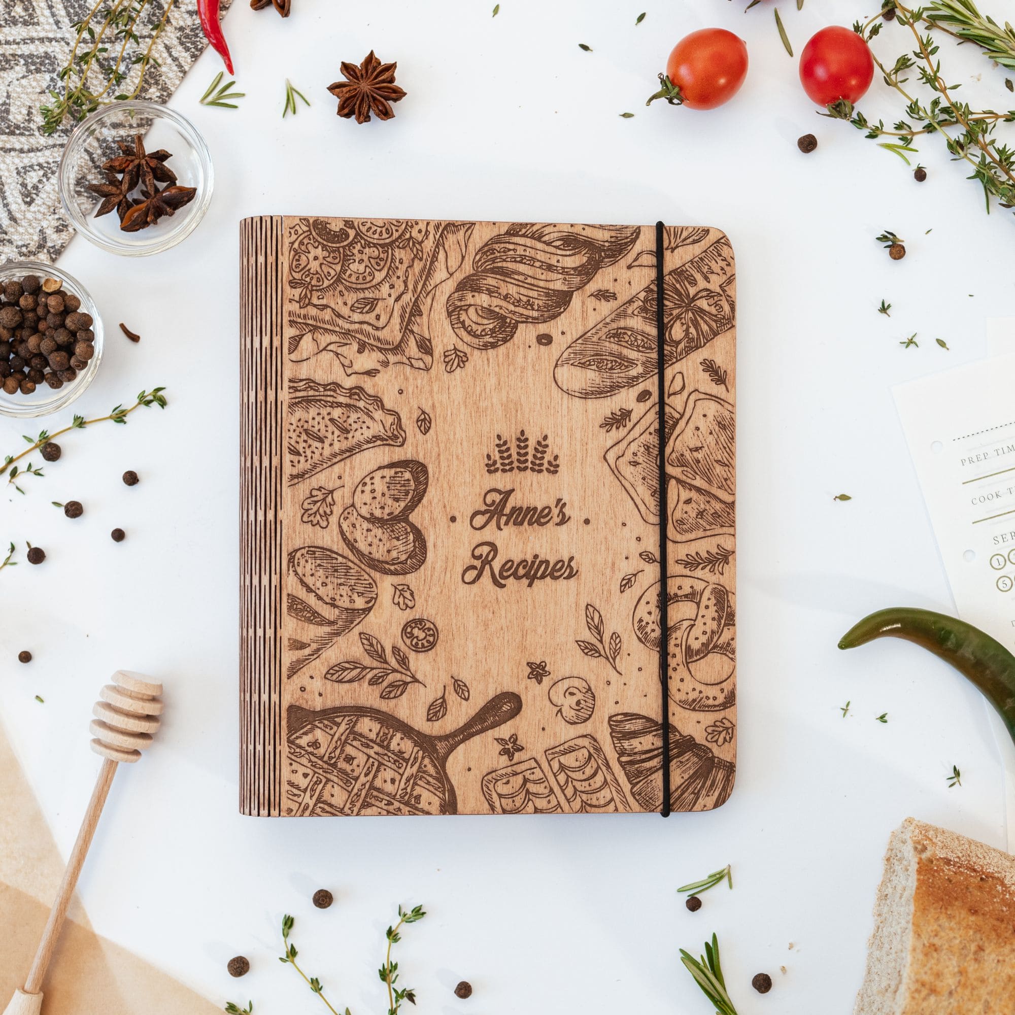 A wooden recipe book with a pretzel design and the text 'Kitchen Recipes' on the cover, surrounded by kitchen-related items like spices and tomatoes.