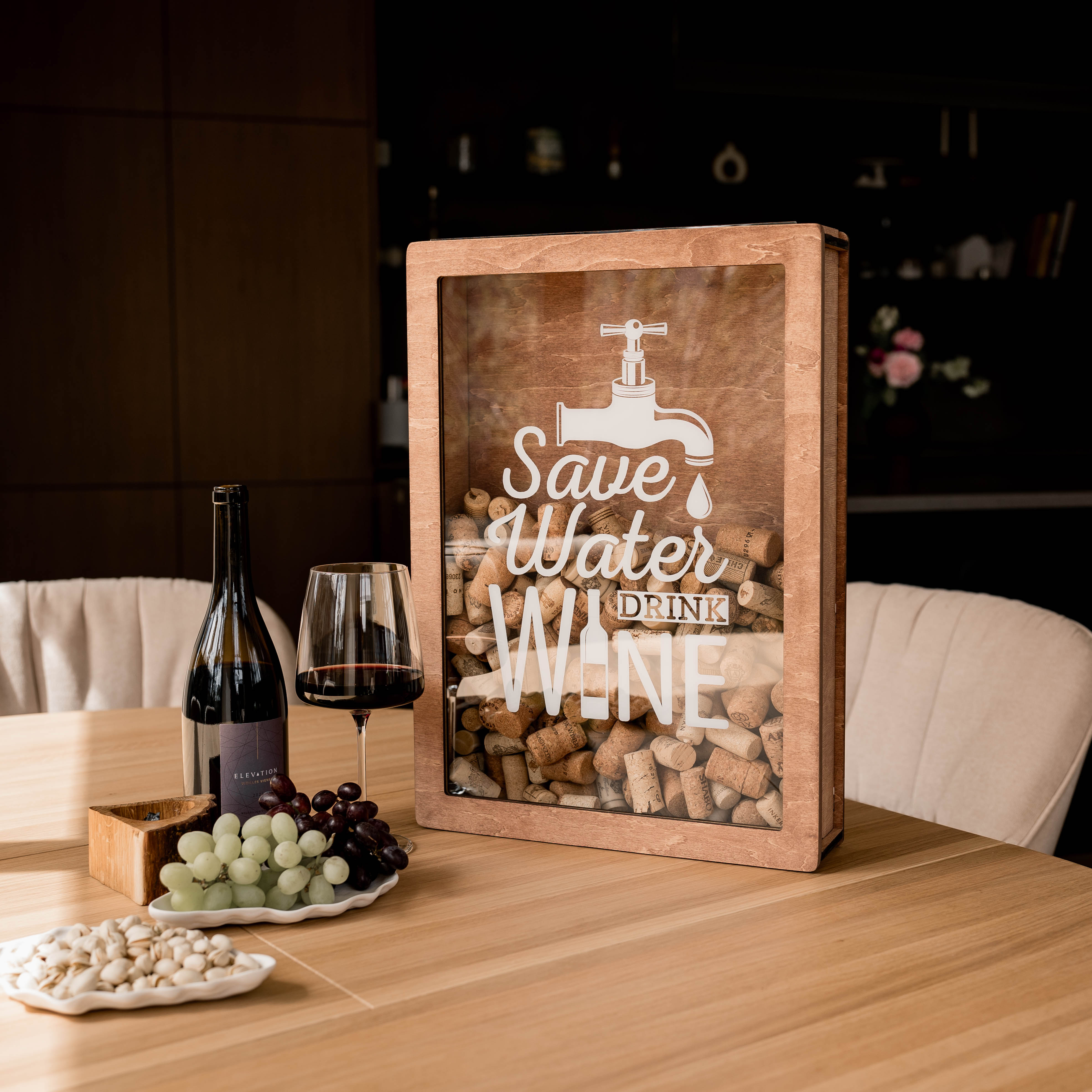 A wooden wine cork box with a glass front displaying text and a wine tap illustration, placed on a table with wine glasses and bottles in the background.