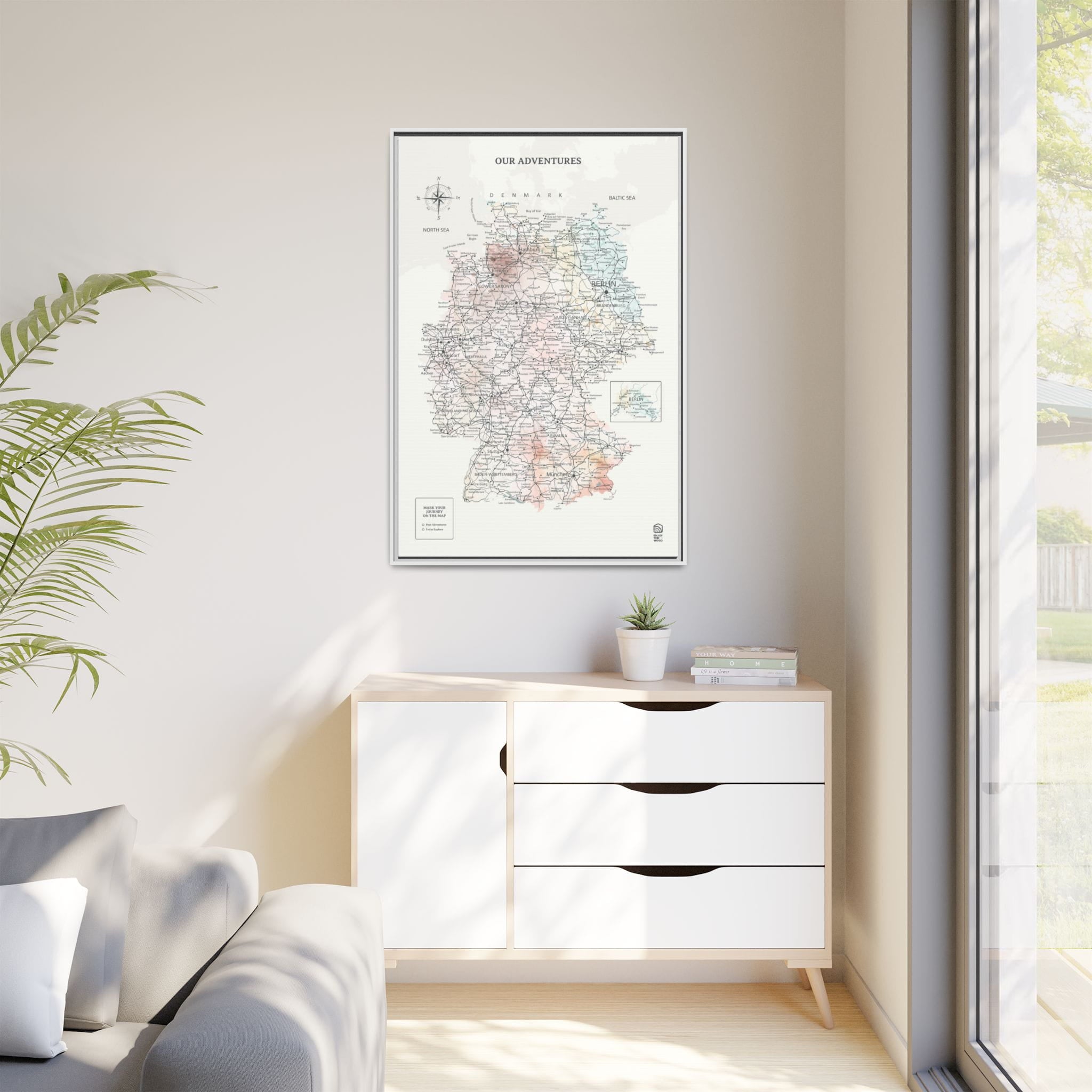 Framed map on a wall above a white cabinet with a plant and books.