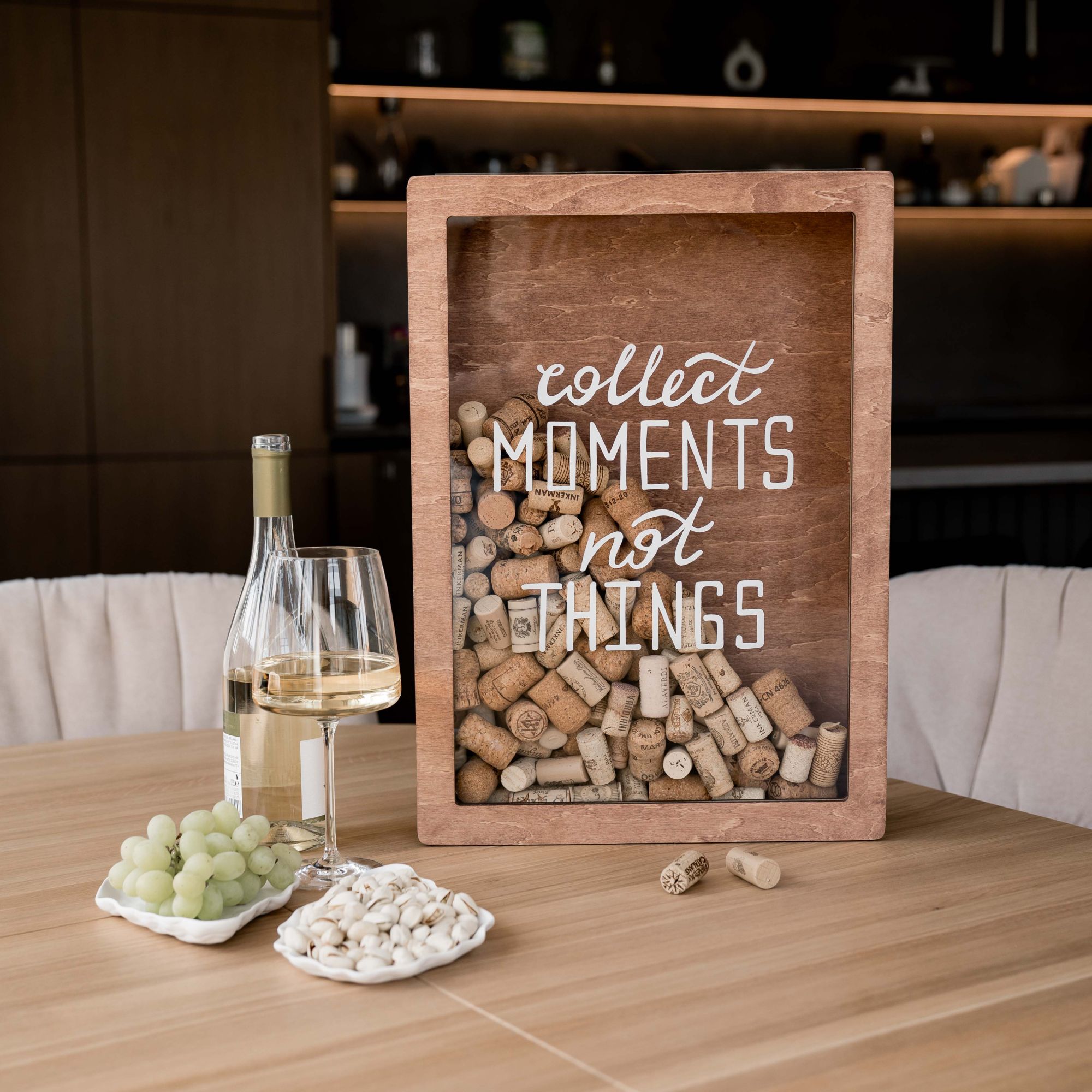 A wooden wine cork box with the text 'collect moments not things' written on the front, placed on a table with a glass of wine and a plate of grapes.