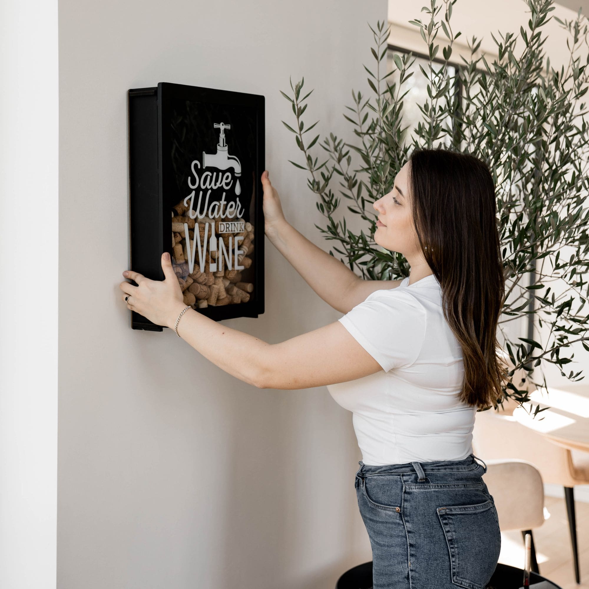 Woman placing cork into black wooden wine cork holder with engraved quote "Save Water Drink Wine", next to white wine bottle, glasses and grapes – modern wine shadow box decor.
