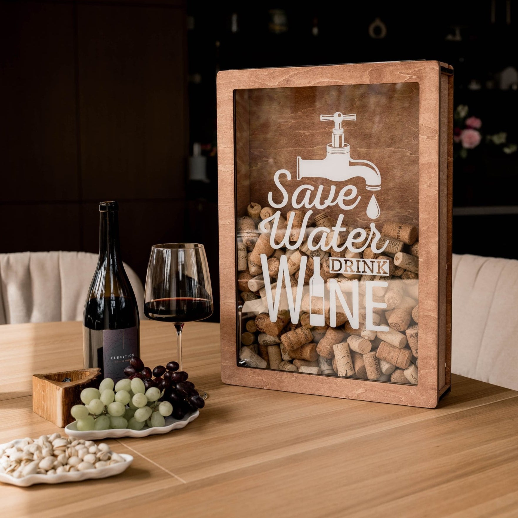 A wooden wine cork box with the text 'Save Water, Drink Wine' displayed on the front, placed on a table with wine glasses and corks visible through the box.