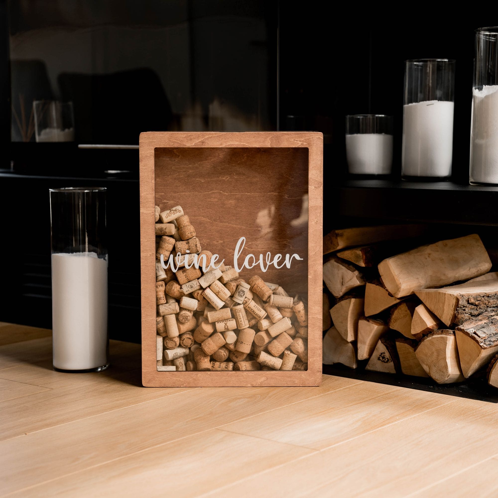 A wooden wine cork box with the text 'Wine Lover' inscribed on it, filled with corks, placed on a table with candles and wood logs in the background.