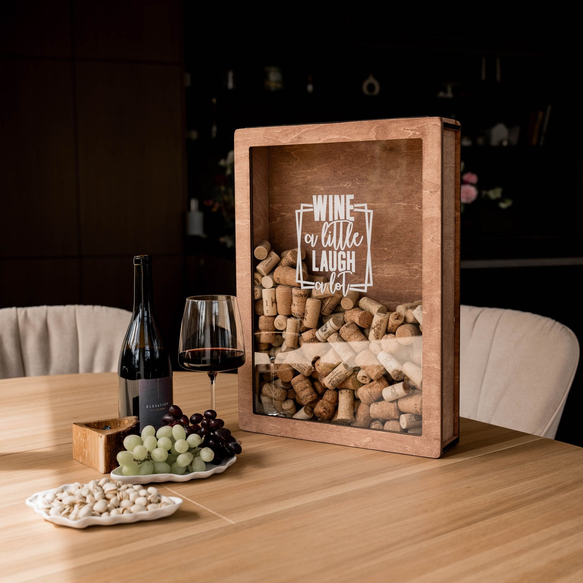 Rustic wooden wine cork shadow box filled with corks, standing on a dining table with wine, grapes, and snacks.