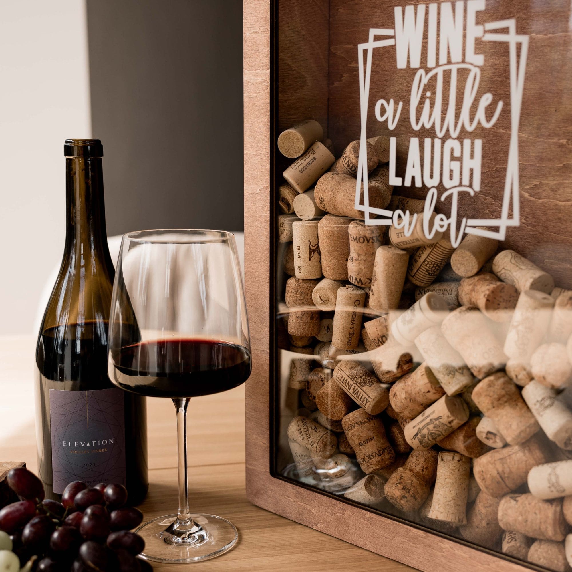 Wooden shadow box for wine corks with the quote "Wine a little, laugh a lot", placed on a table next to a wine bottle, glass of red wine, and grapes.