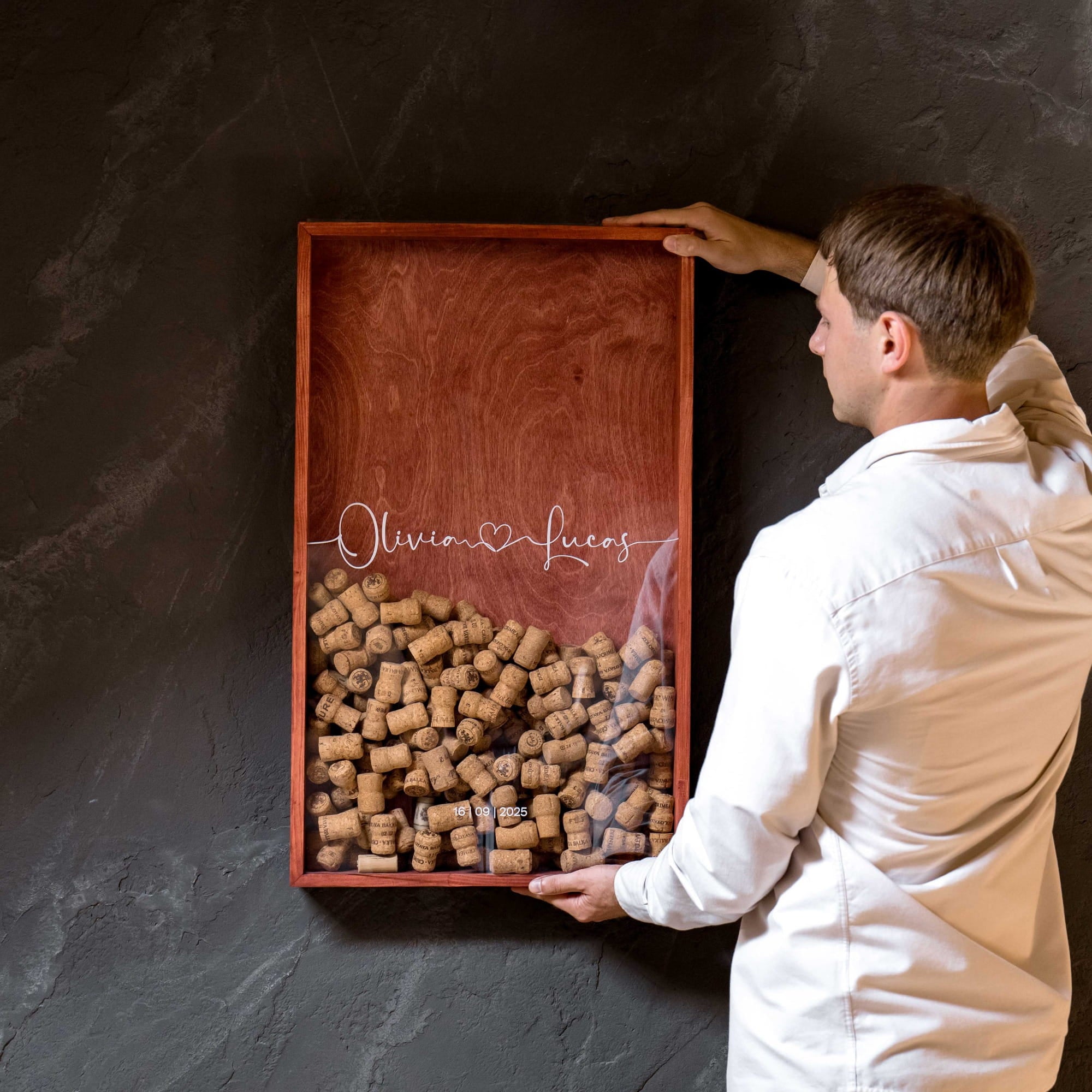 A man in a white shirt holds a custom wooden wine cork tray mounted on a dark textured wall