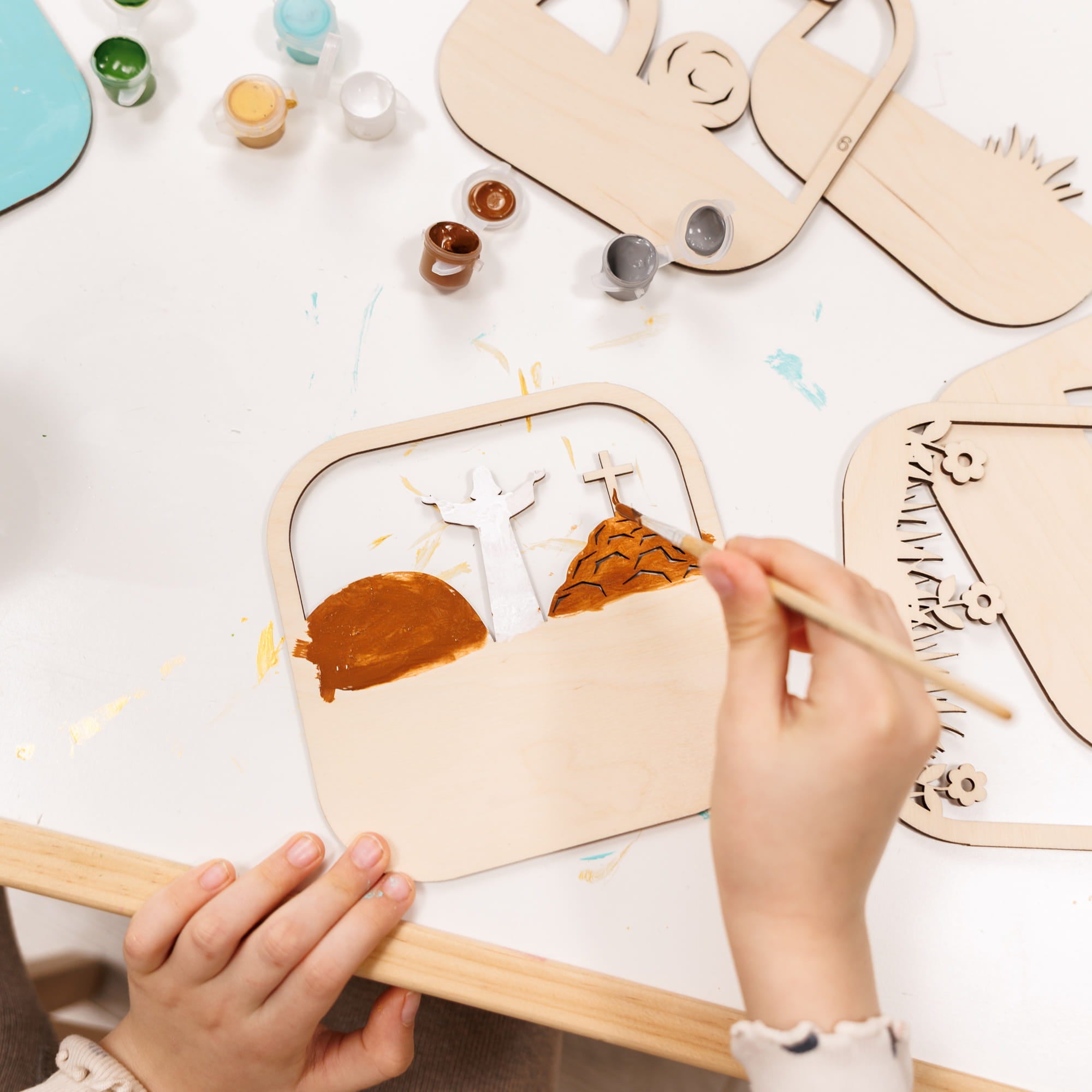 Children's hands painting wooden animal shapes on a white surface with paint bottles and brushes.