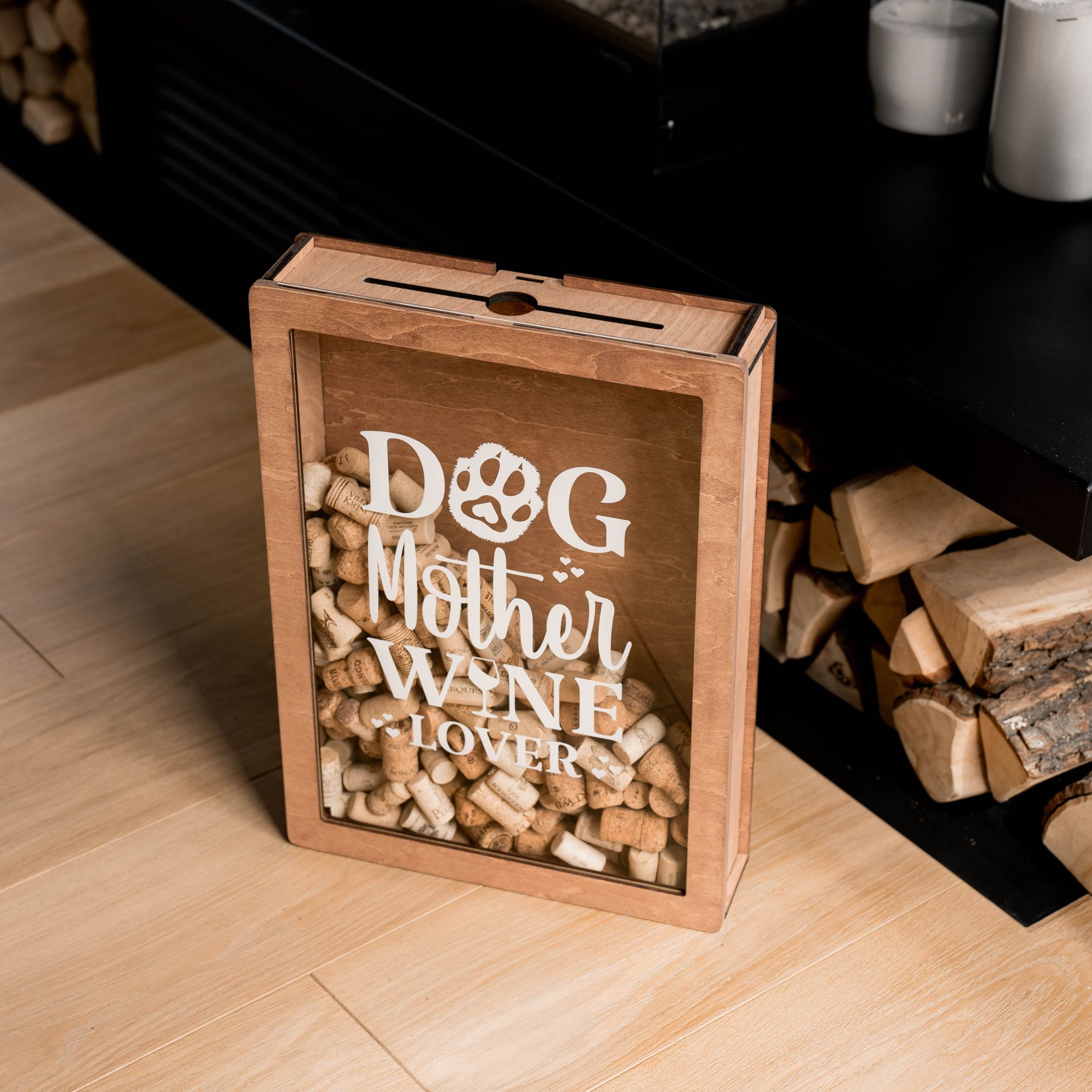 A wooden cork box with the inscription 'Dog Mother Wine Lover' on the front, filled with wine corks, and placed on a floor beside a wooden bench.