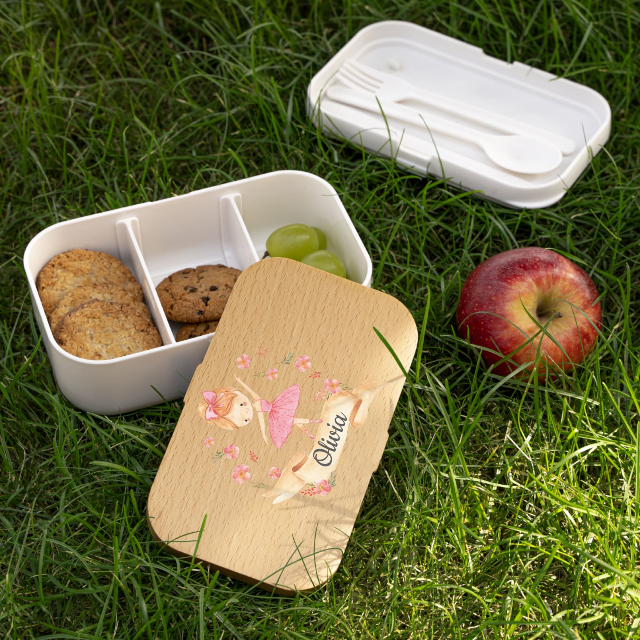 White bento box with compartments on grass, containing cookies and grapes, with a wooden lid featuring a fairy design.