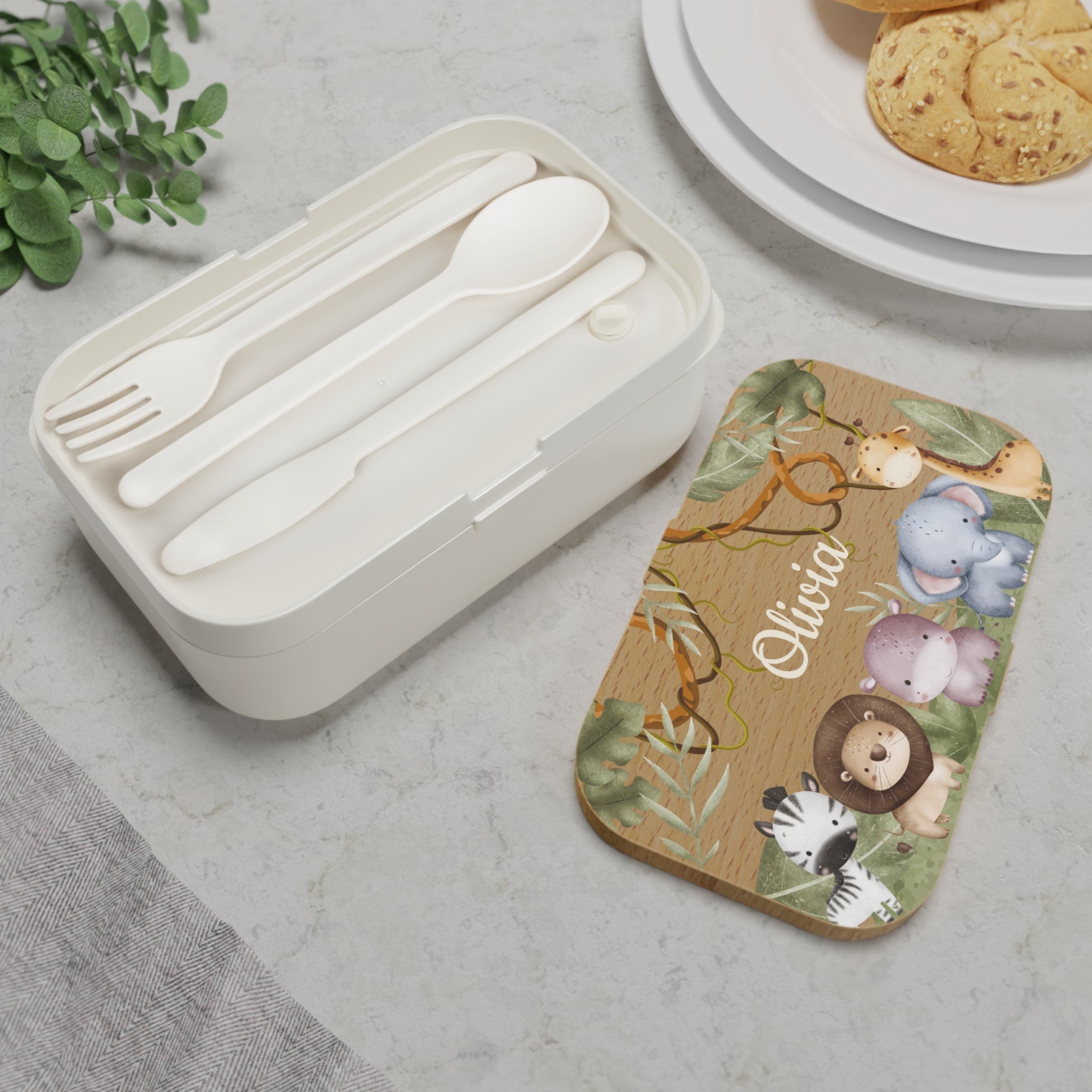 Set of white cutlery in a container next to a wooden coaster with animal designs and the name 'Olivia'.