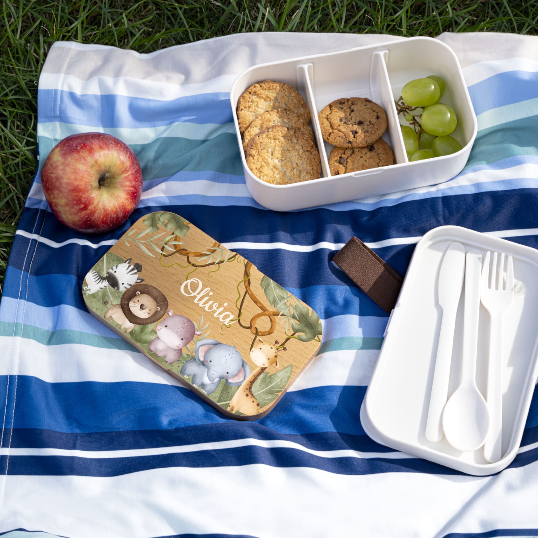 Picnic setup with a bento box, apples, cookies, and utensils on a striped blanket outdoors.