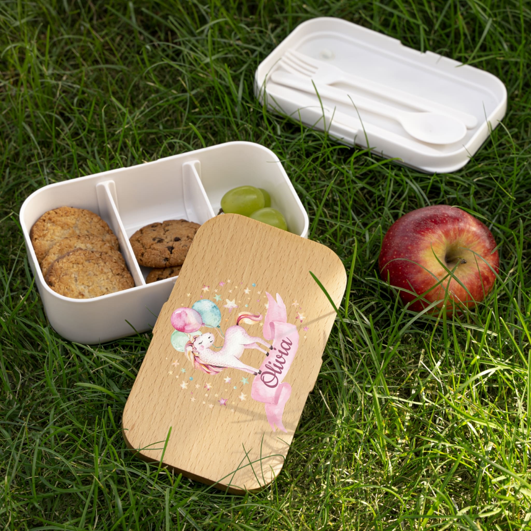 Bento box with compartments, cookies, grapes, and an apple on grass with a wooden nameplate featuring a unicorn design.