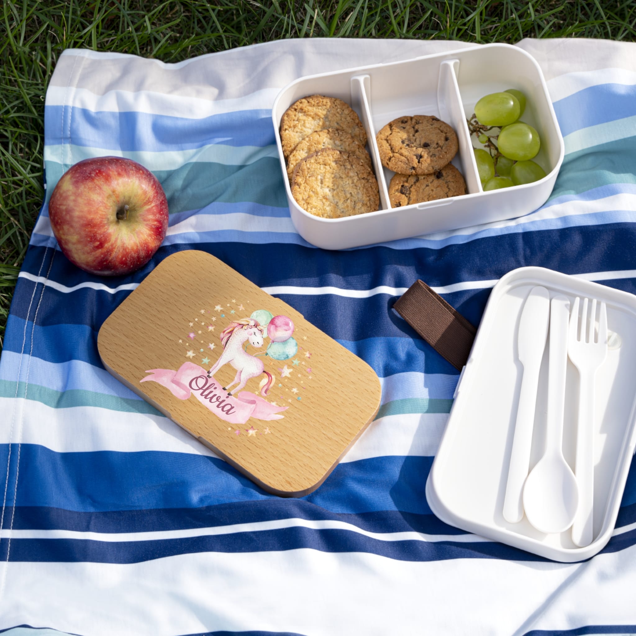 Picnic setup with a personalized wooden tray, food container, and snacks on a striped blanket outdoors.