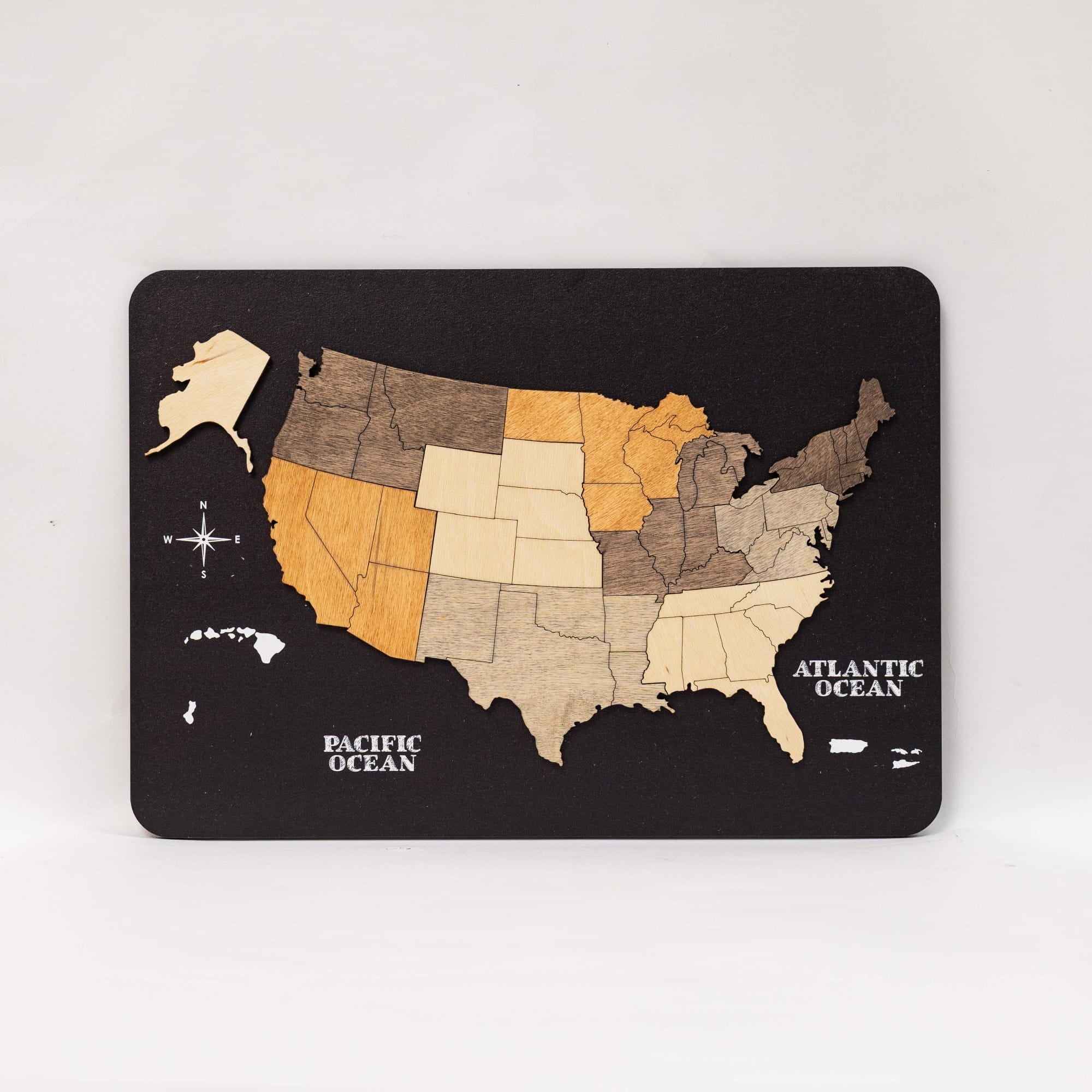 A rectangular black plaque featuring a wooden USA map with states in varying natural wood tones, labeled with Pacific Ocean, Atlantic Ocean, and a small compass rose, photographed on a light gray background.