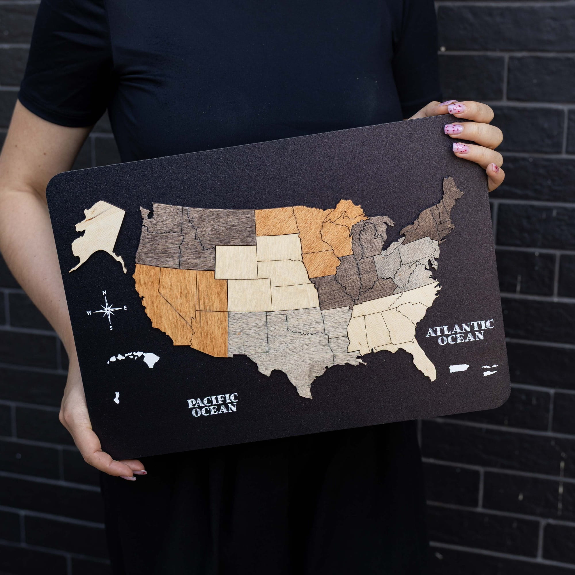A person holds a rectangular black plaque featuring a wood-grain USA map with states in light beige, gray, and orange-brown tones, plus labeled oceans. The person is wearing a black top and standing against a dark brick wall.