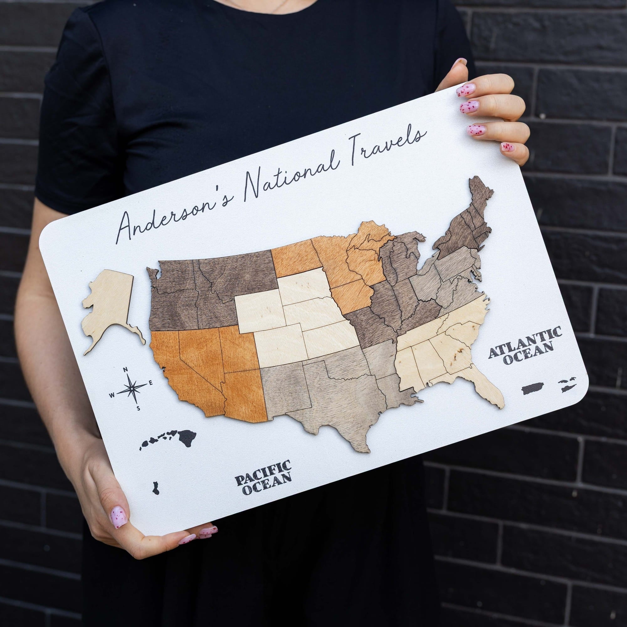 A person in a black shirt holds a white rectangular wooden plaque featuring a stylized map of the United States labeled 'Anderson's National Travels' with 'Pacific Ocean' and 'Atlantic Ocean' text. The person's hands and torso are visible against a dark brick wall.