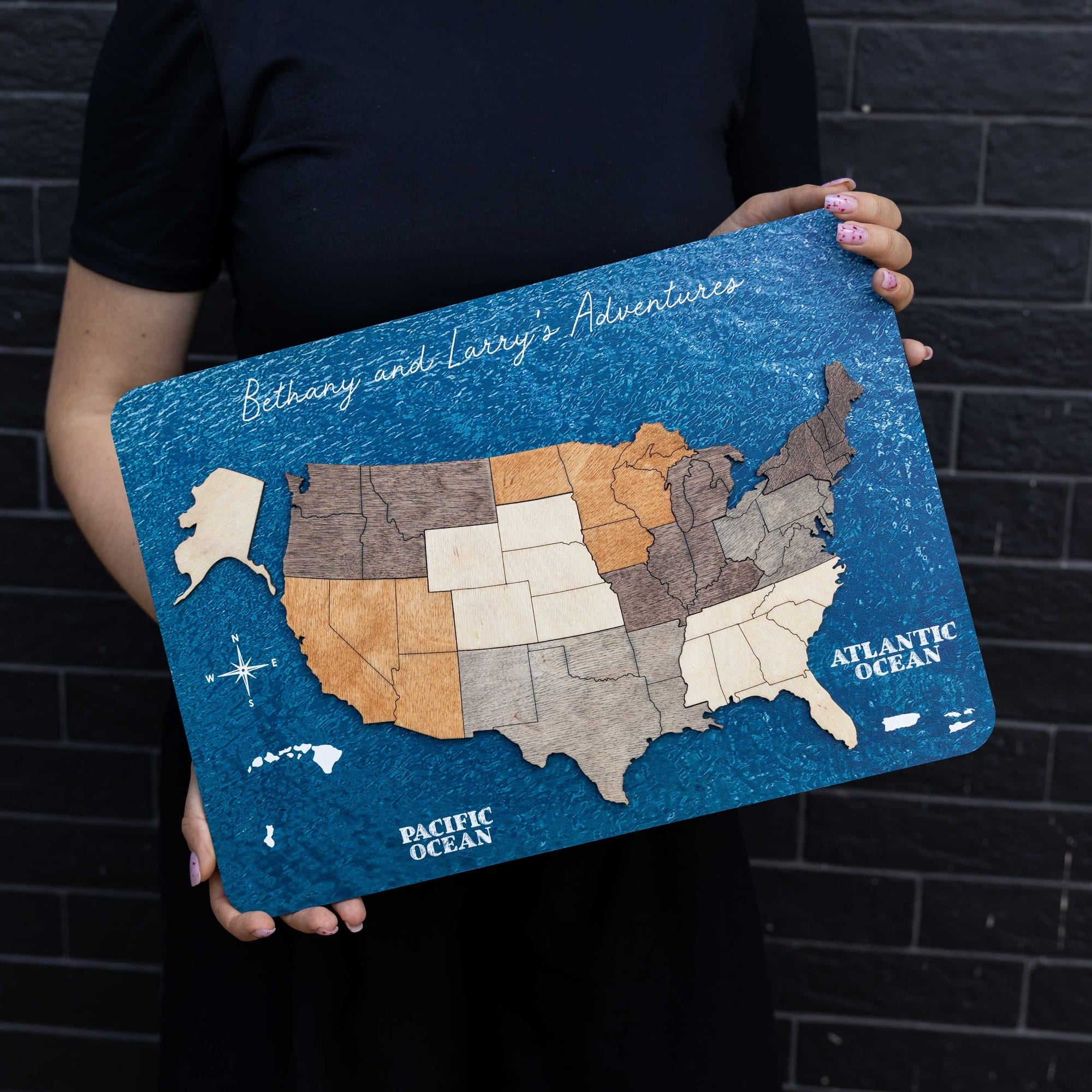 A person holds a rectangular wooden US map board featuring a textured blue ocean design, state outlines, and the text 'Bethany and Larry's Adventure' at the top. The background is a dark gray brick wall.