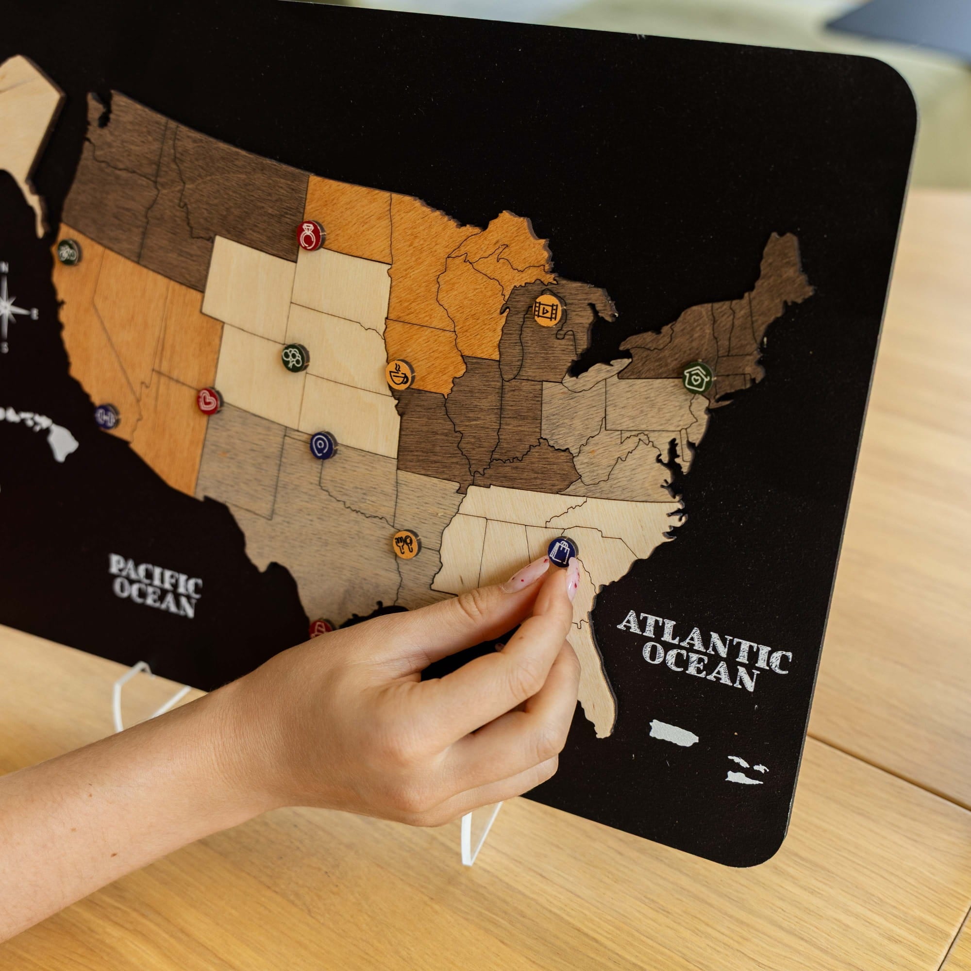 A close-up of a hand placing a small map pin on a black wooden United States travel-style map board with state sections in different wood tones, photographed on a light wood tabletop.
