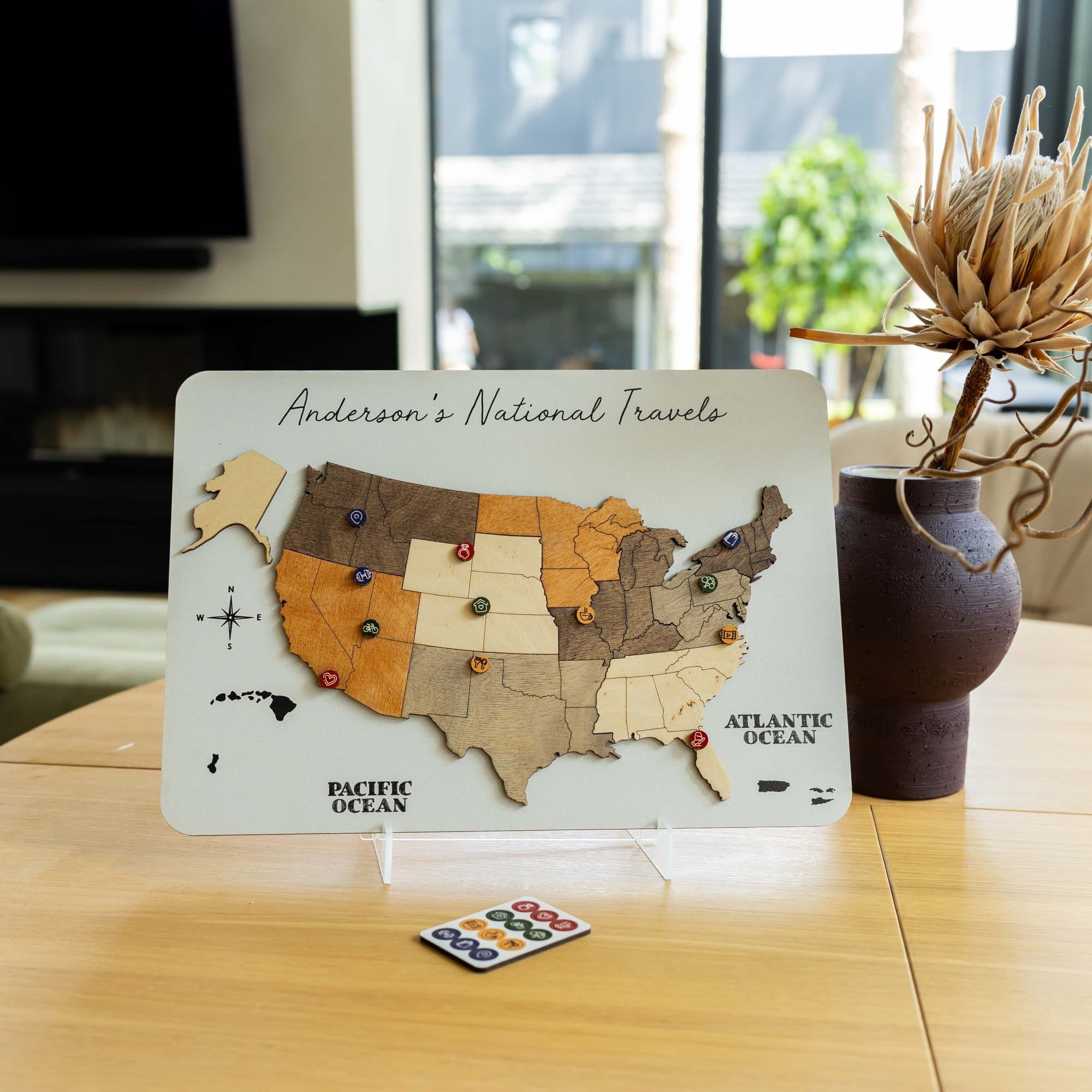 A wooden United States travel map plaque on a clear acrylic stand, displayed on a wooden table in a home setting next to a vase with dried flowers.