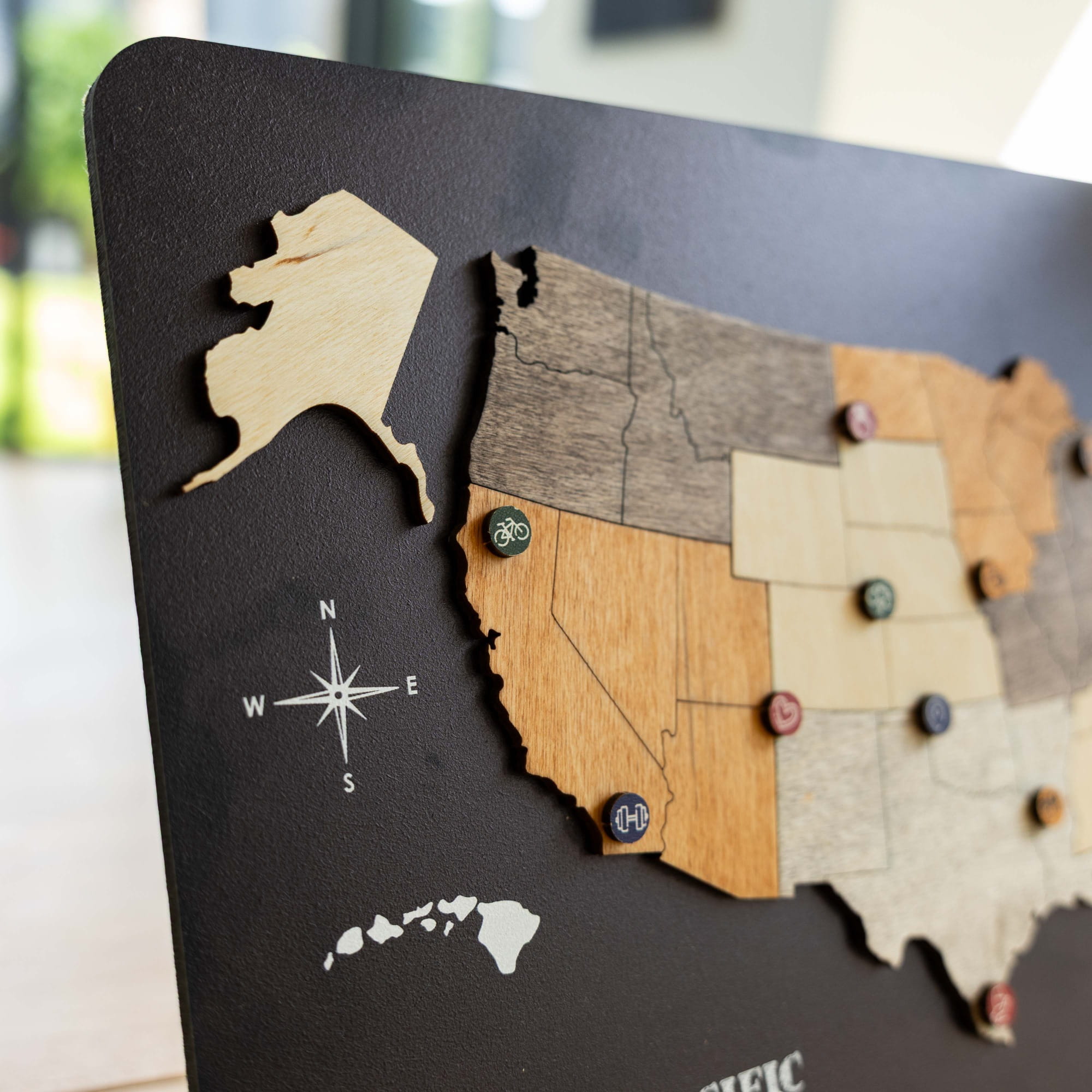 A close-up of a wooden US state map plaque on a dark background, with Alaska, California, Oregon, Washington, and Hawaii shown in detail. The map uses different wood tones and small colored pin markers.