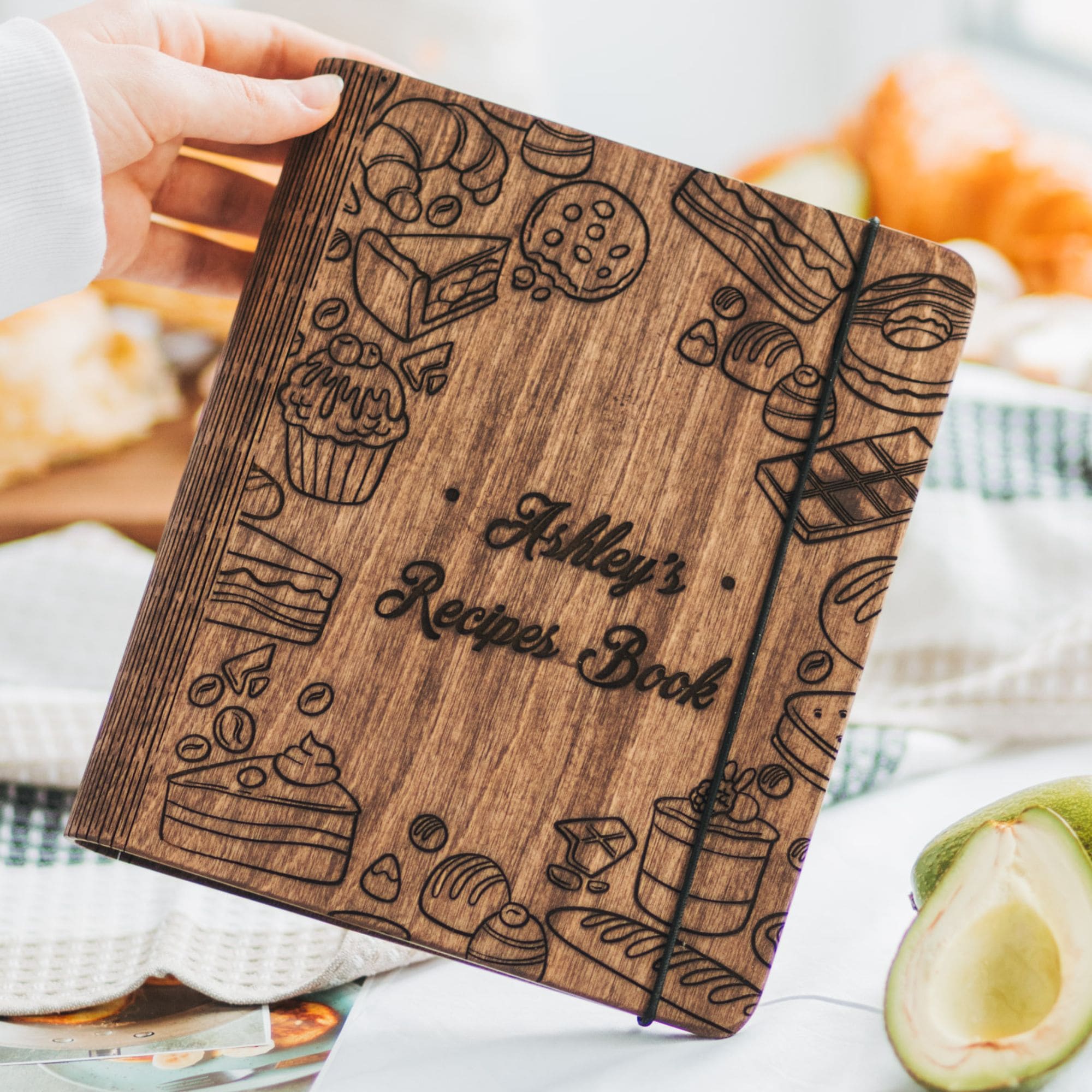 Wooden recipe book engraved with "Ashley’s Recipes Book," held above a white towel with avocado and bread in the background.
