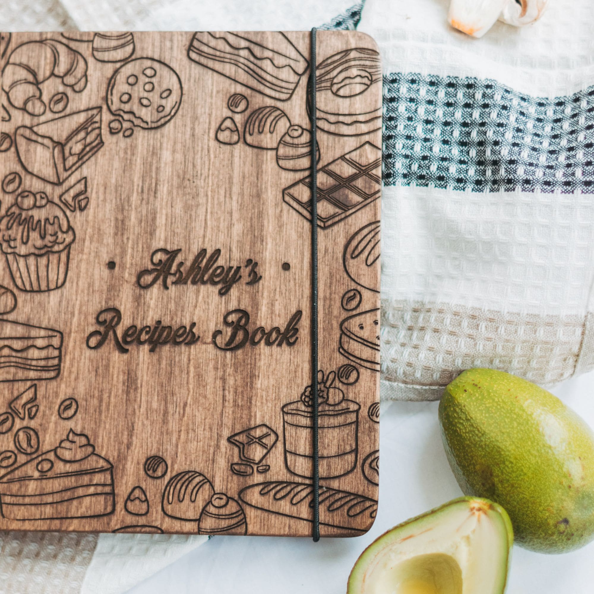 Personalized wooden recipe book with engraving "Ashley’s Recipes Book" placed on a towel with fresh avocado.