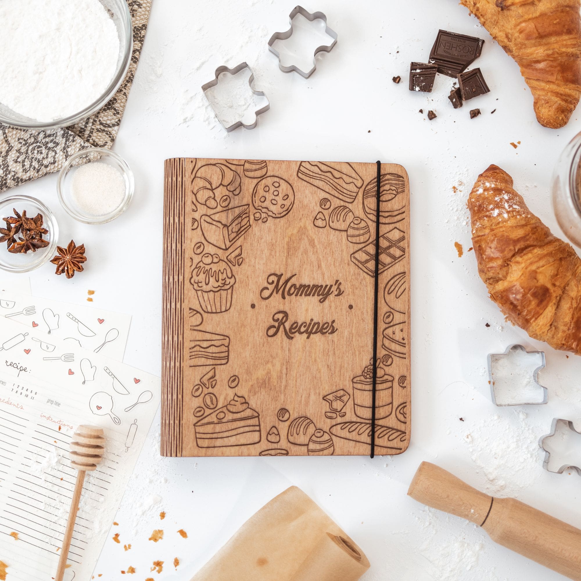 A wooden recipe book with a pastries design, titled 'Mommy's Recipes', surrounded by baking-related items like chocolate pieces, a honey dipper, and baking decorations on a white background.
