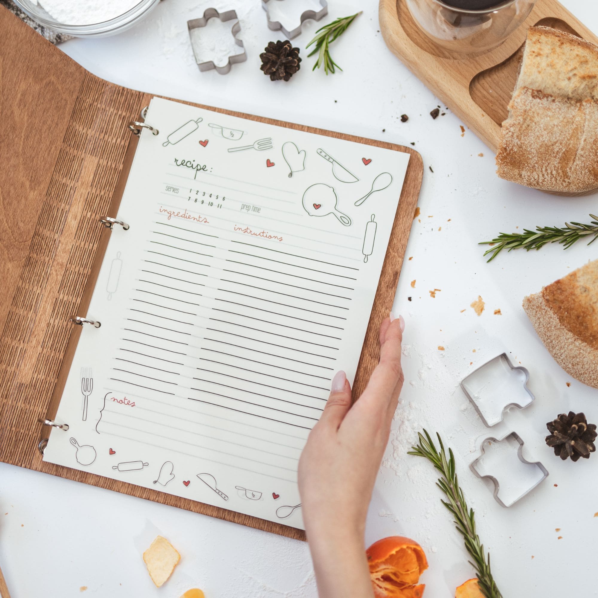 Open wooden recipe binder with illustrated recipe page, surrounded by baking tools, bread, and herbs.