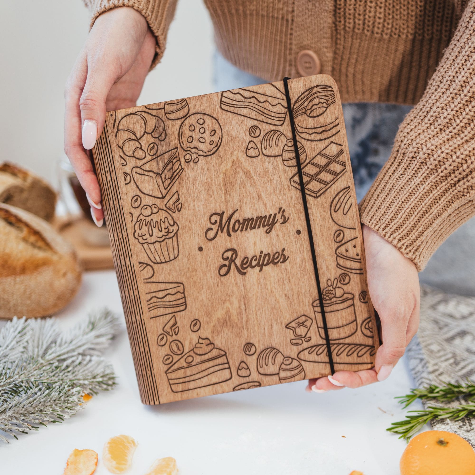 Person holding wooden engraved recipe book "Mommy’s Recipes," surrounded by bread, greenery, and citrus on the table.