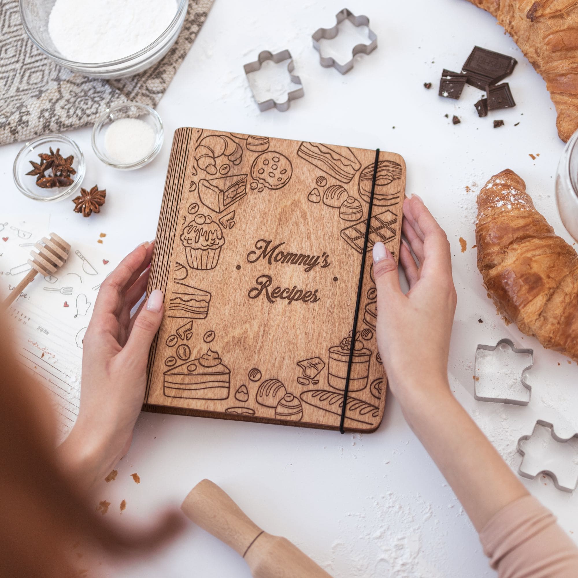 Wooden recipe book with engraving "Mommy’s Recipes" surrounded by baking ingredients and croissant on a white table.