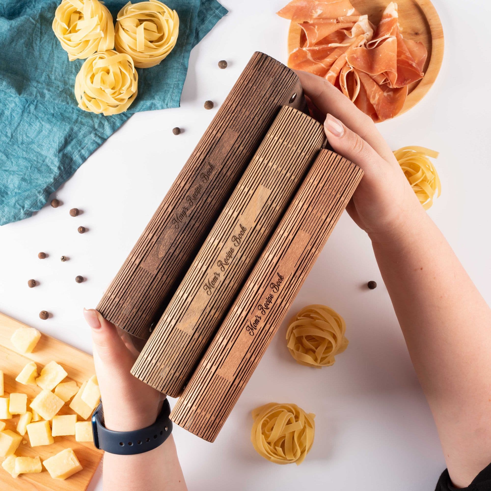 Three engraved wooden recipe books in different shades held in hands, with pasta and cheese on the table.