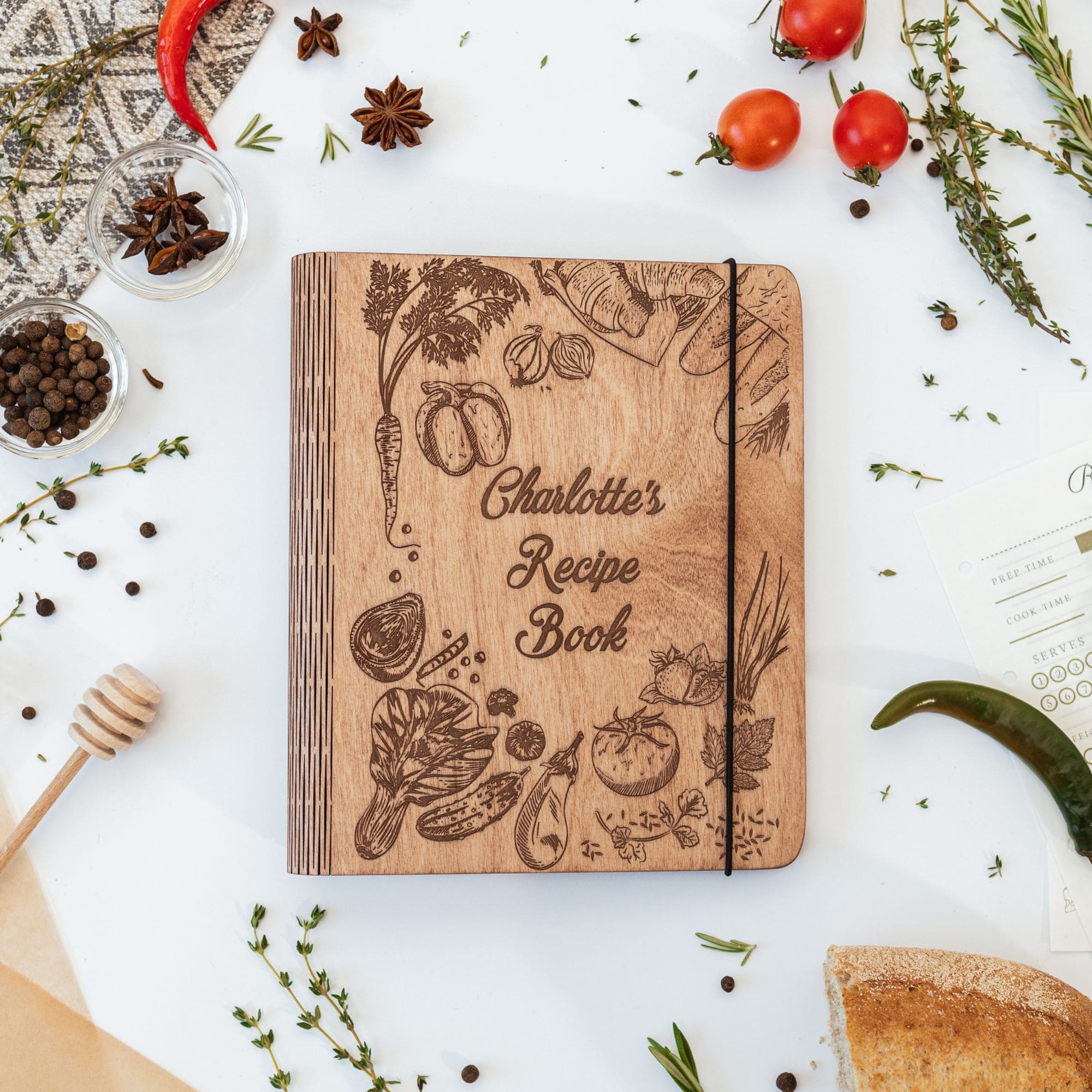 A wooden recipe book with a peppers theme cover, personalized with the name 'Charlotte's Recipe Book'. The cover is adorned with engraved illustrations of vegetables, fruits, and spices.