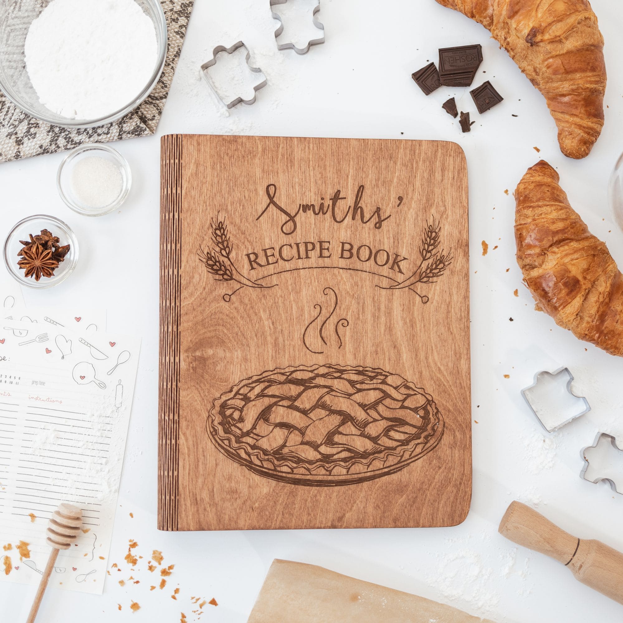 A wooden recipe book with a pie design on the cover, surrounded by baking ingredients and utensils on a white background.