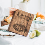 A wooden recipe book with a pie design on the cover, personalized with the name 'Rachel'. The book is displayed on a table with a pie and fruits in the background, indicating it may be used for recording family recipes.