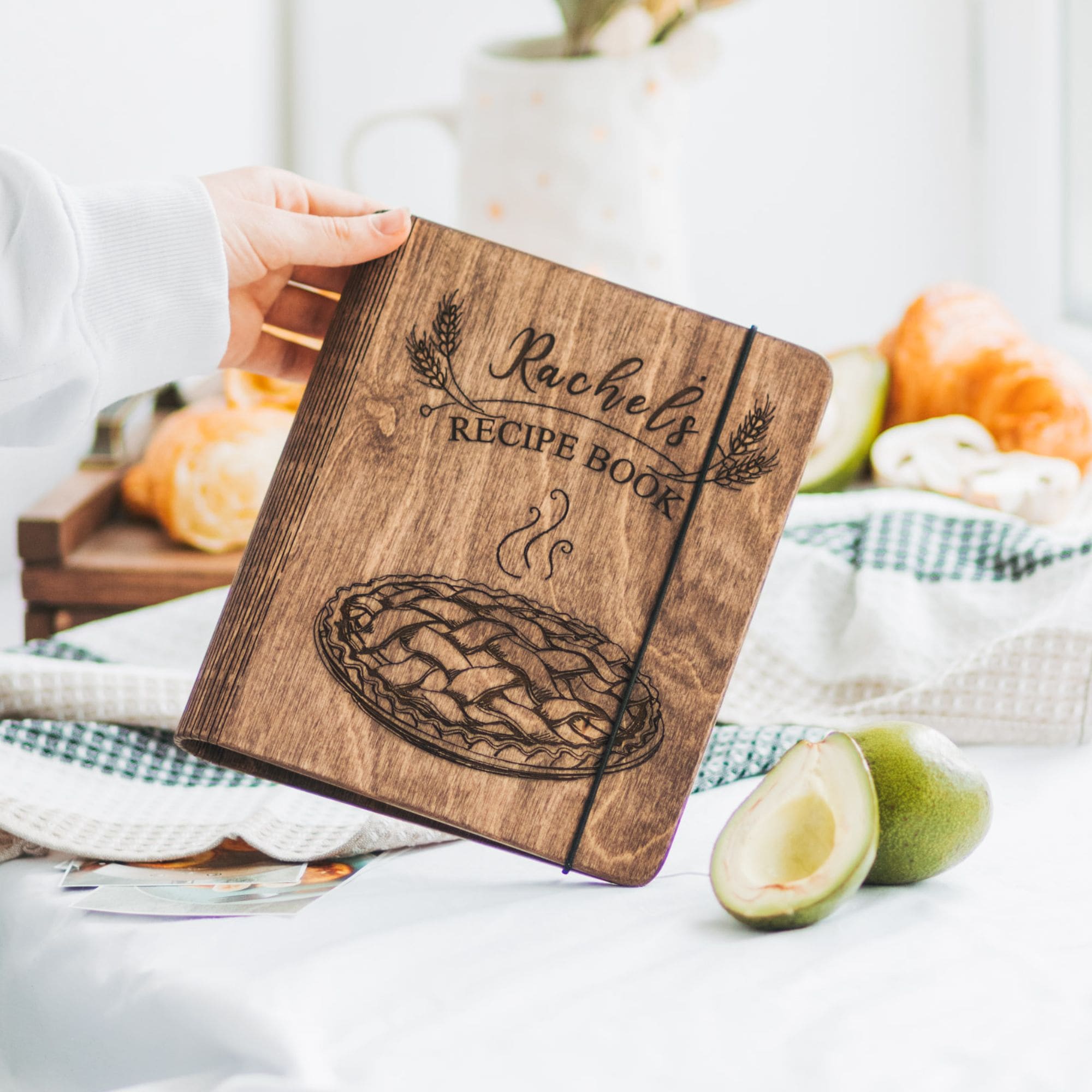 A wooden recipe book with a pie design on the cover, personalized with the name 'Rachel'. The book is displayed on a table with a pie and fruits in the background, indicating it may be used for recording family recipes.