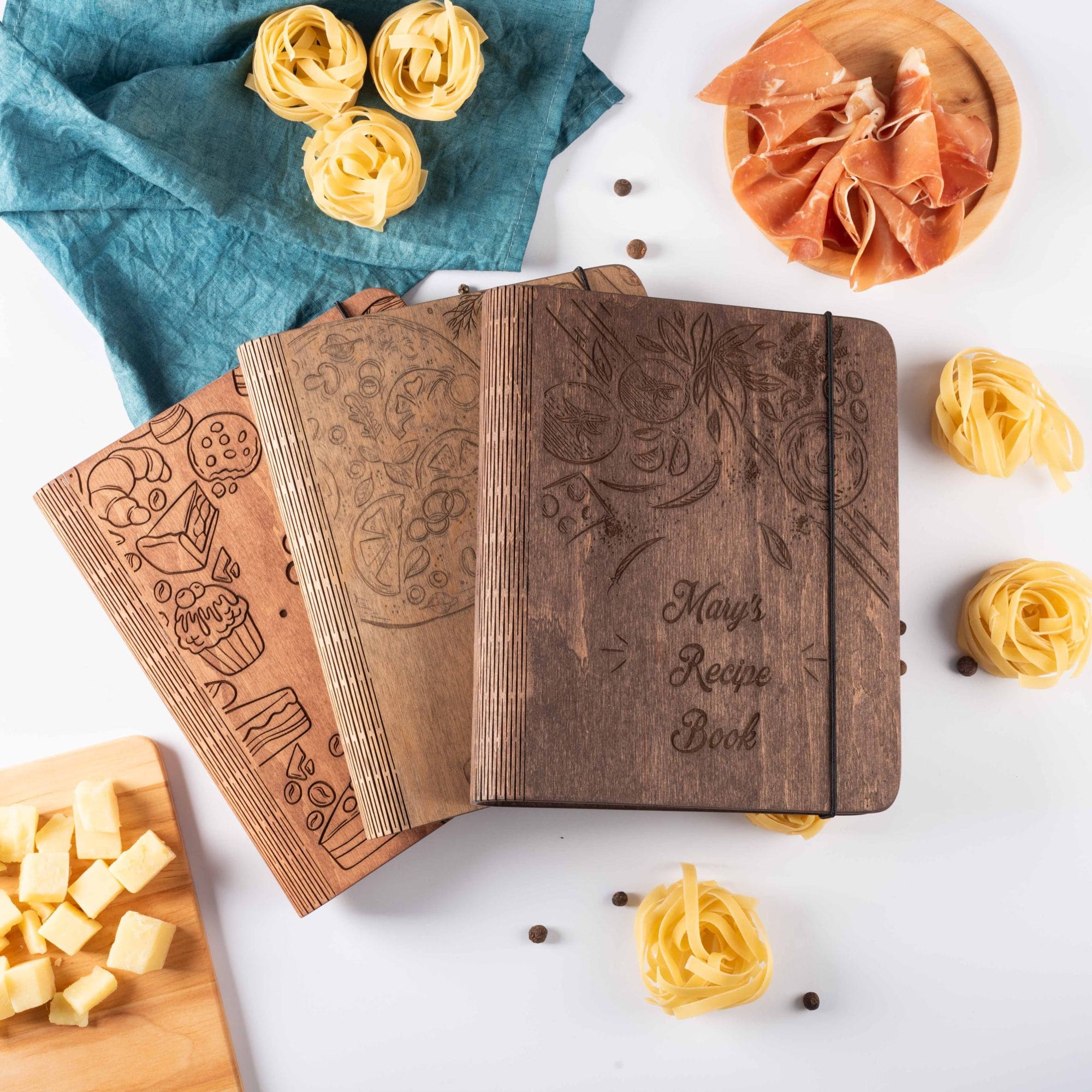 Set of three engraved wooden recipe books with different designs, surrounded by pasta and cheese.