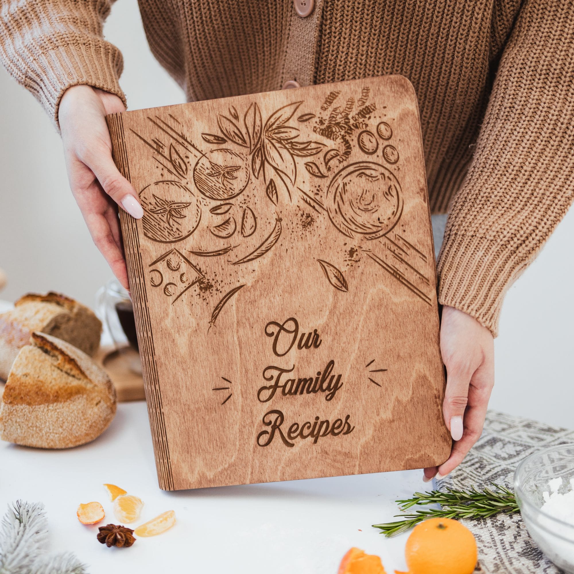 A person holding a wooden recipe book with a floral design and the title 'Our Family Recipes' on the cover.
