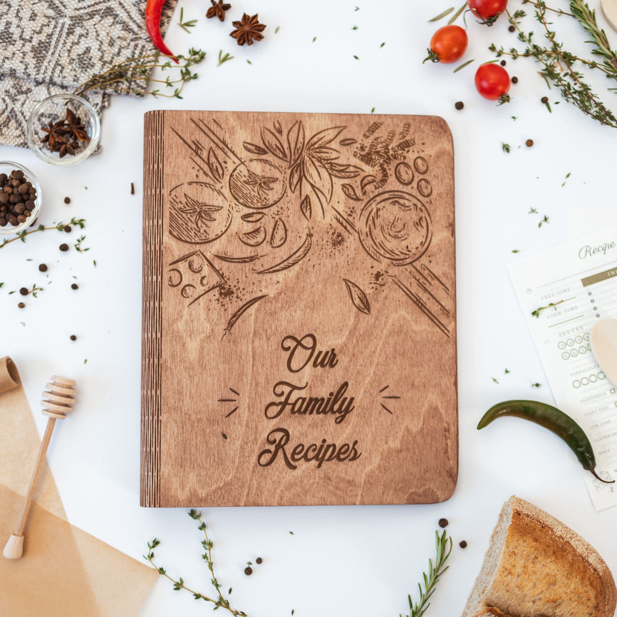 A wooden recipe book with a tomatoes theme, titled 'Our Family Recipes', surrounded by food items and a honey dipper on a white background.