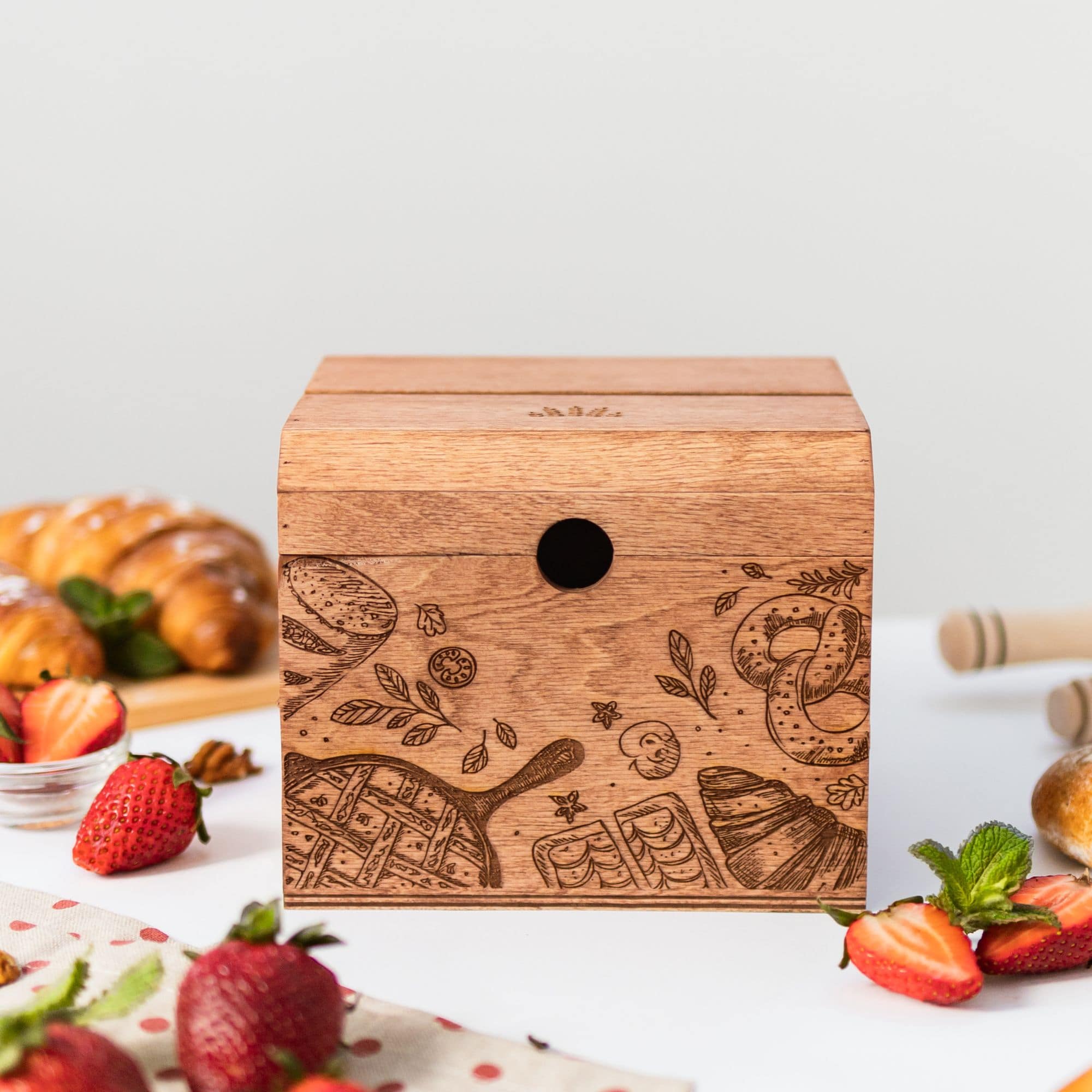A wooden recipe box with a bakery theme, featuring pretzel designs and utensils, placed on a kitchen counter surrounded by strawberries, bread, and a notebook.