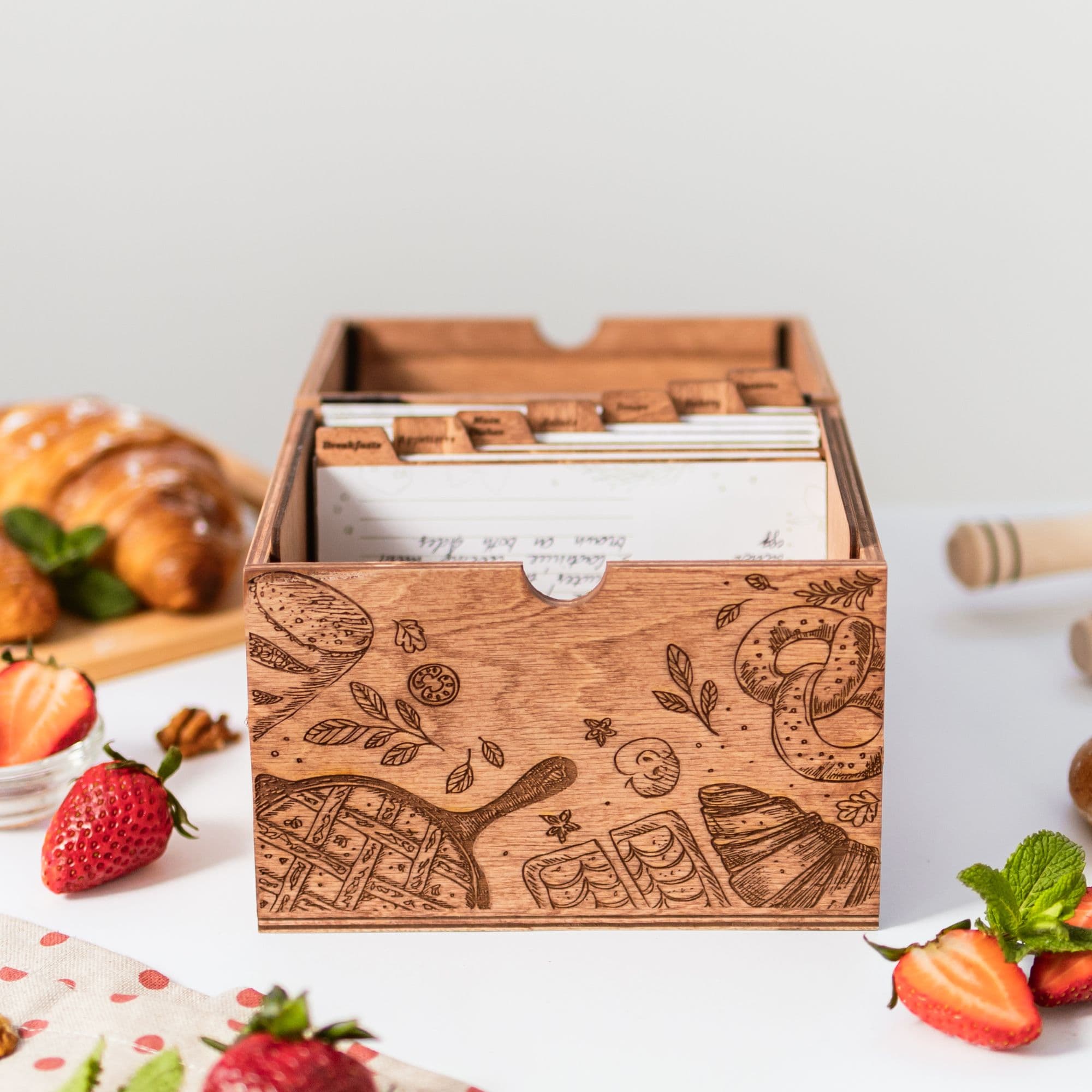 Wooden recipe box with engraved kitchen design and recipe cards inside, surrounded by strawberries and croissants