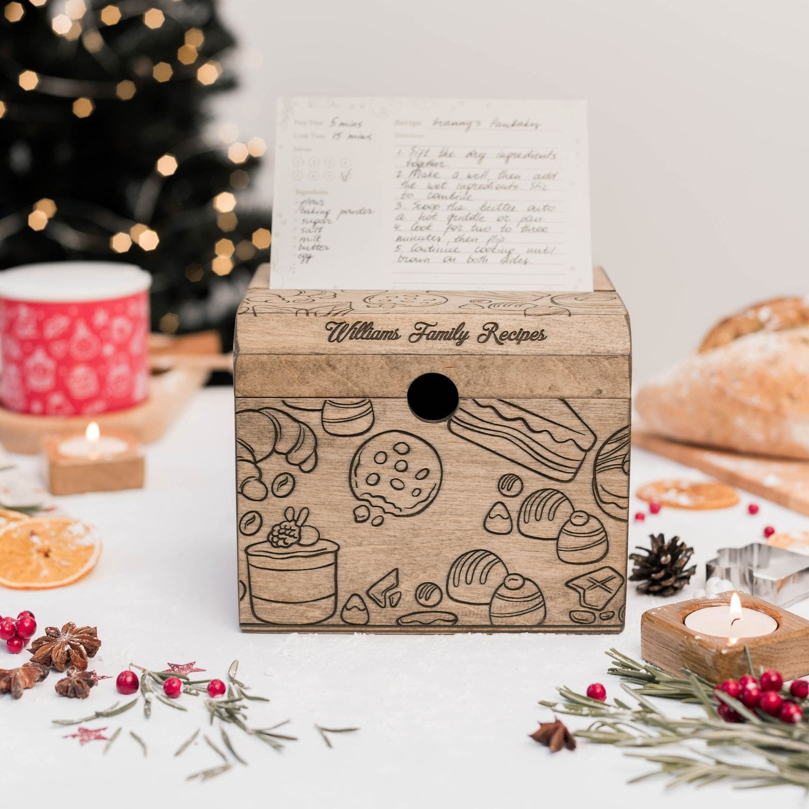A wooden recipe box with pastries artwork displayed on a table with festive decorations, including candles and pine cones, with a recipe card partially inserted into the box.