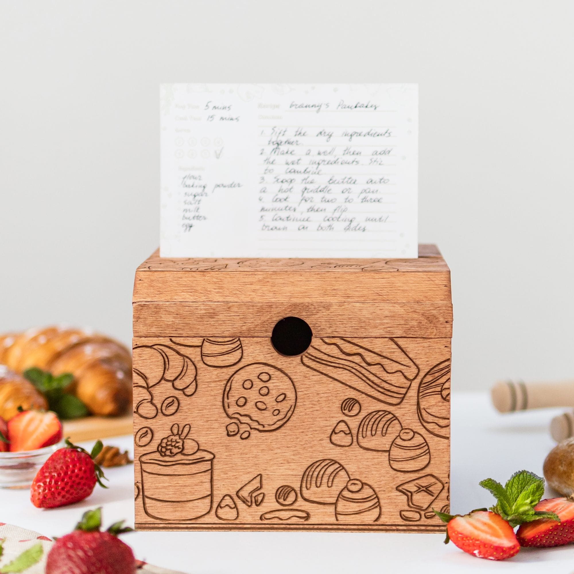 Engraved wooden recipe box with handwritten recipe card on top and strawberries around