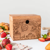 A wooden recipe box with pastries collection, decorated with pastry artwork, placed on a kitchen counter surrounded by strawberries, a honey dipper, and other kitchen items.