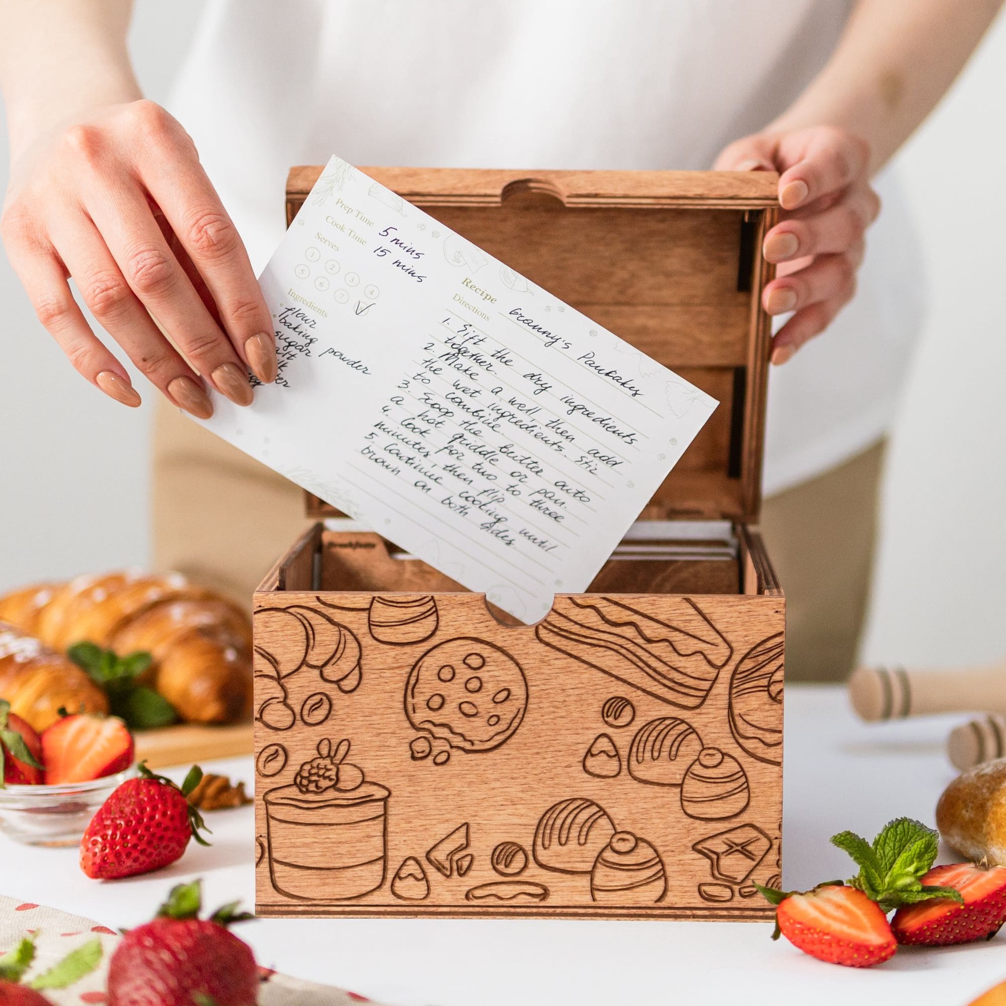 A person is in the process of writing on a recipe card with a pen. The card is placed in a wooden box that has a whimsical design with illustrations of food items.