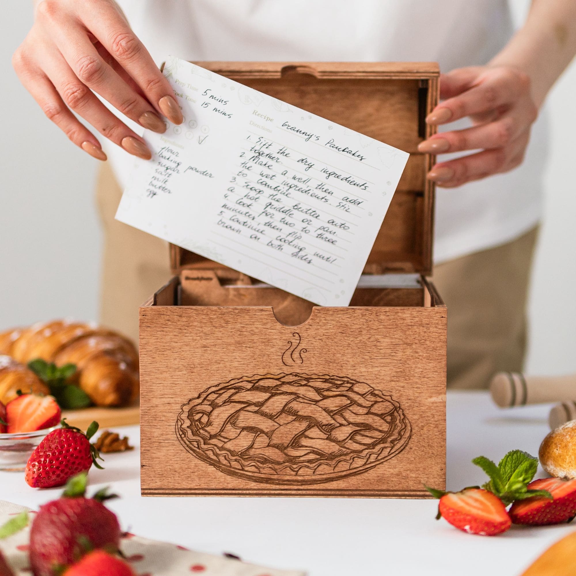 Wooden recipe box with engraved pie design, person inserting recipe card.