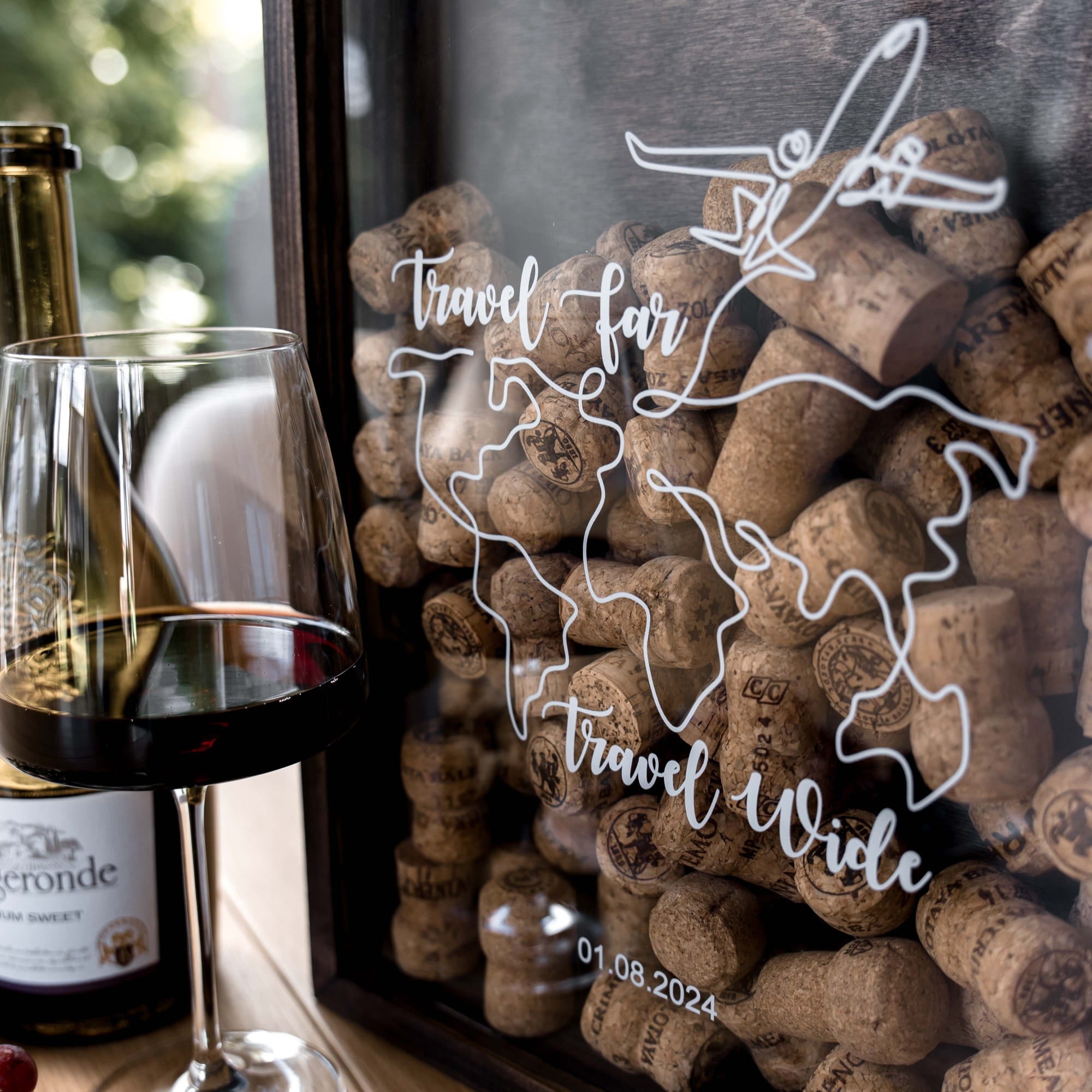 A close-up of a personalized wine bottle or glass container filled with cork stoppers, featuring white line art of a globe and the words 'travel far travel wide' on the front.