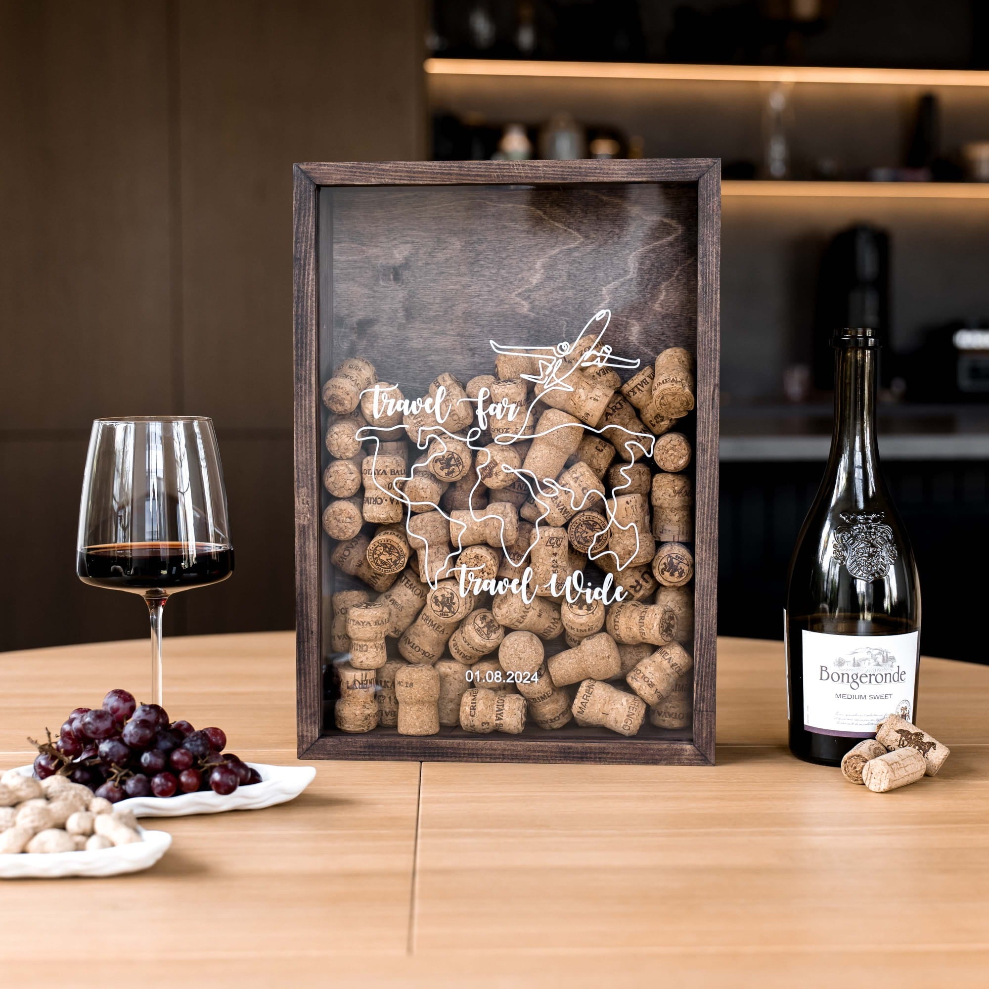 A framed wine-themed cork board display on a wooden table, surrounded by a glass of red wine, a bottle of Bourgogne wine, and plates of grapes and nuts.
