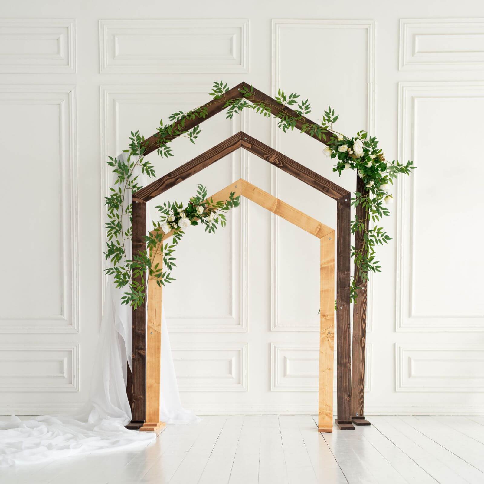 A pentagon-shaped wooden wedding arch decorated with greenery, set against a white background.